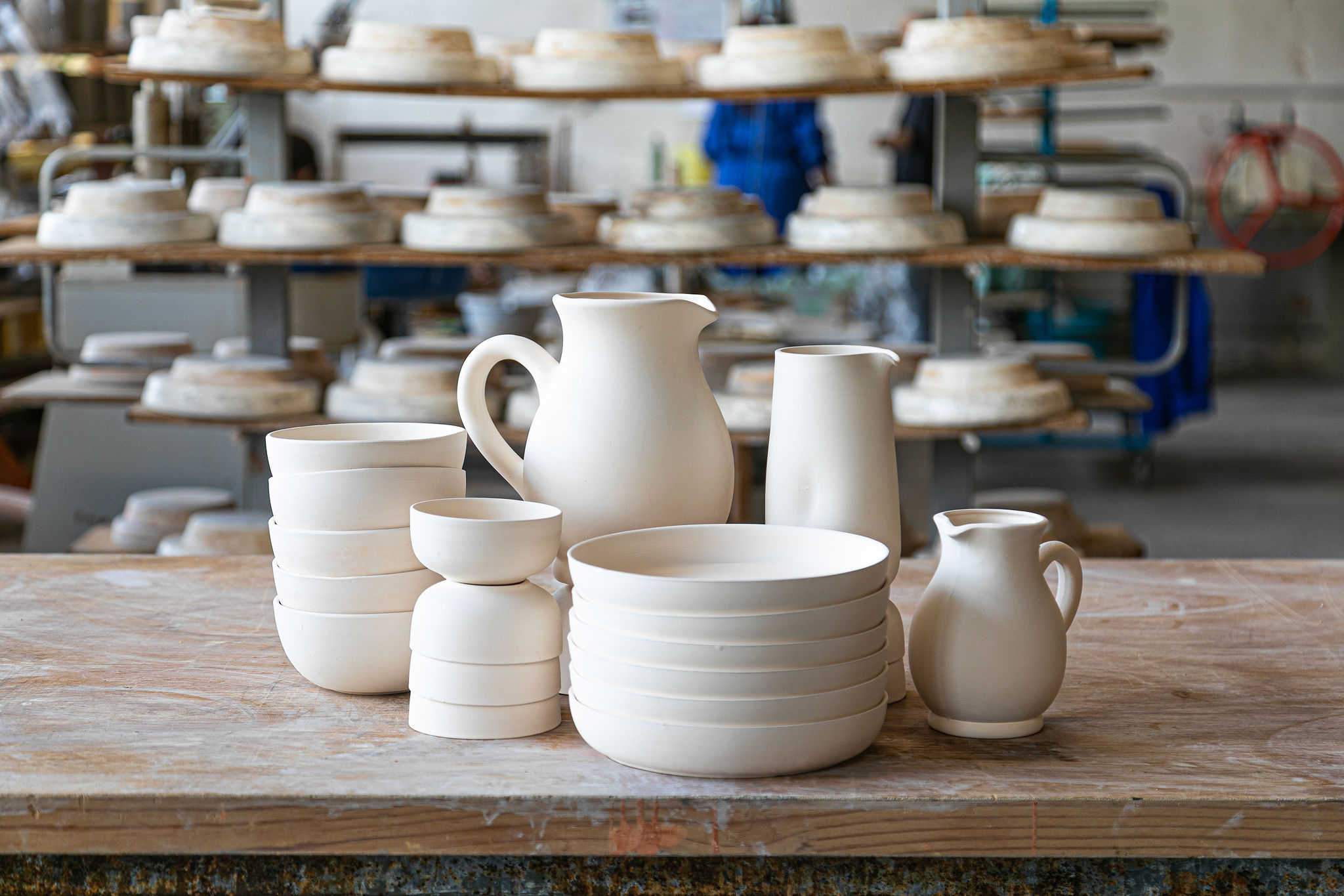 Des pièces de céramique blanches non cuites (pichets, bols, assiettes) sont arrangées sur une table en bois, dans un atelier de poterie rempli d'étagères d'autres pièces.