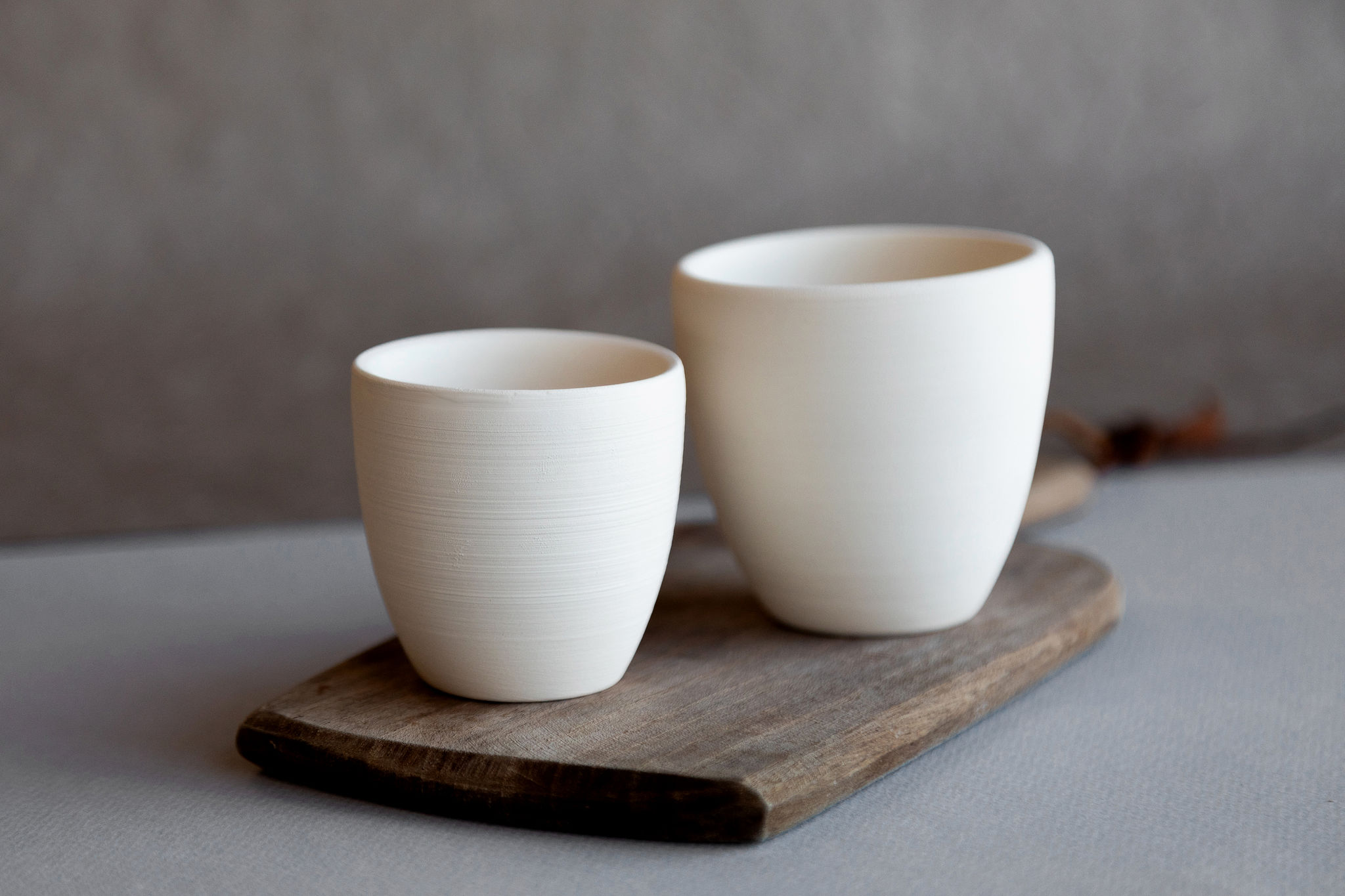 Deux tasses en céramique blanches, l'une plus petite, sont posées côte à côte sur une planche en bois rustique. Le fond est un mur gris flou.
