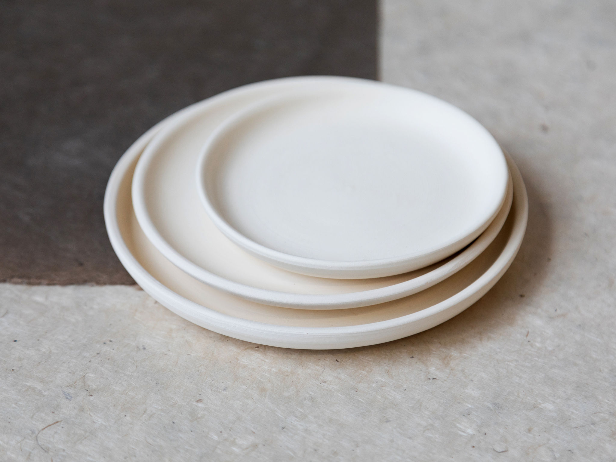 Trois assiettes rondes en céramique blanche mate sont empilées par taille décroissante, reposant sur une surface claire et texturée avec un arrière-plan flou plus foncé.