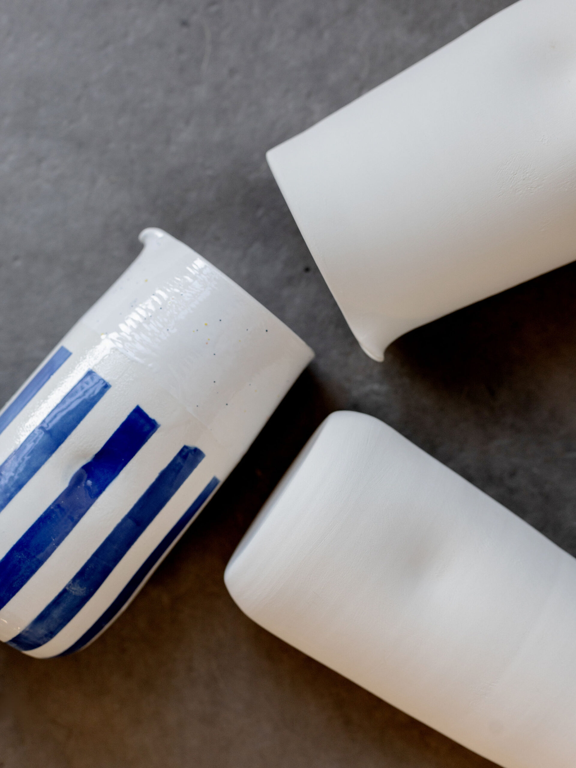 Trois tasses en céramique, deux blanches et une rayée bleu et blanc, sont posées en diagonale sur une surface gris foncé, vues de dessus.