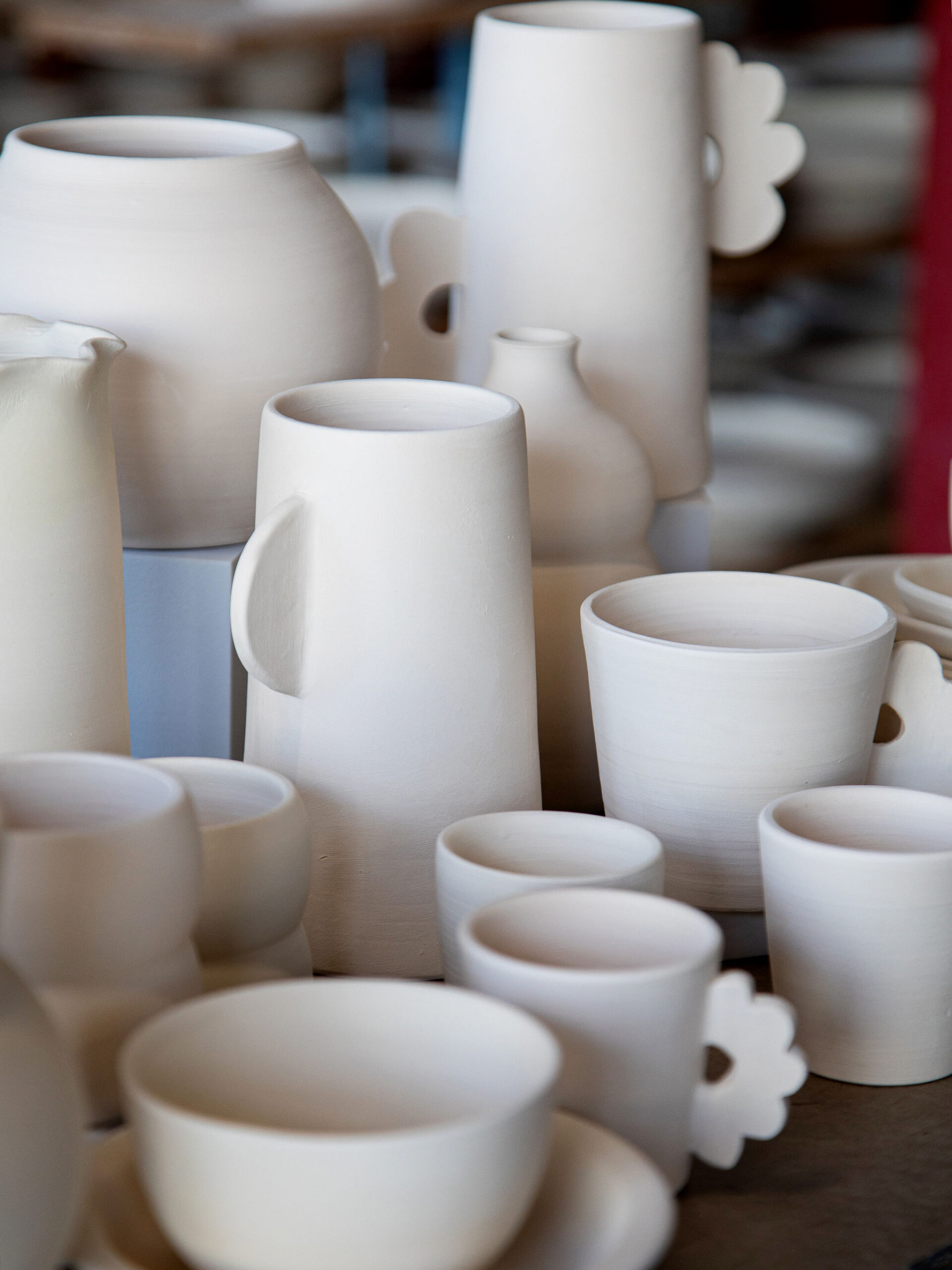 De nombreuses pièces de céramique blanche non émaillée, incluant tasses et vases avec certaines anses florales, sont présentées dans un environnement d'atelier flou.
