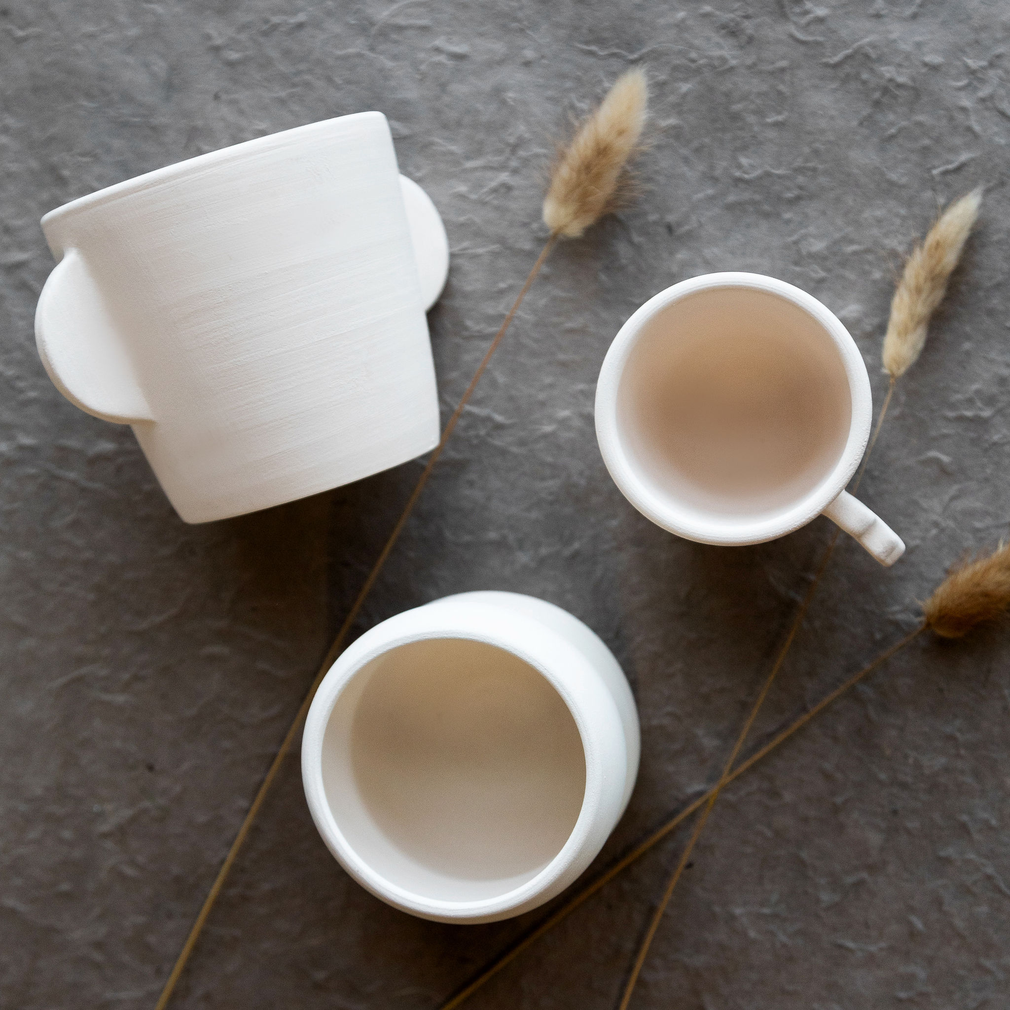 Trois tasses claires, dont une tasse d'apprentissage à deux anses et deux petites tasses, sont posées sur une surface texturée gris foncé, agrémentée d'herbes séchées.