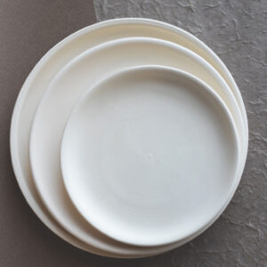 Trois assiettes rondes blanches s'empilent du plus grand au plus petit. Elles reposent sur une surface bicolore, gris foncé lisse à gauche et gris clair texturé à droite.