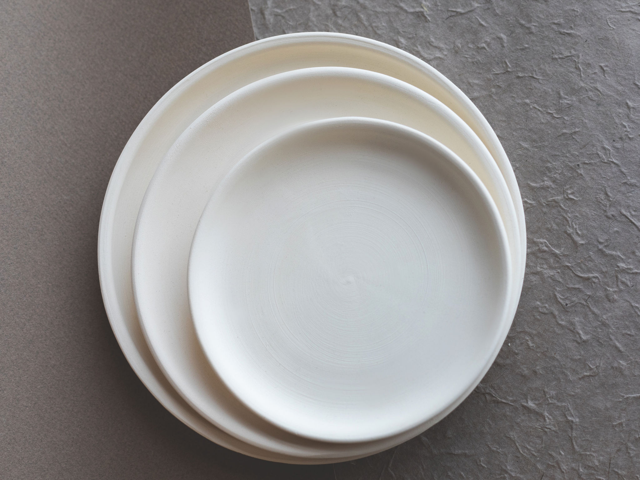 Trois assiettes rondes blanches s'empilent du plus grand au plus petit. Elles reposent sur une surface bicolore, gris foncé lisse à gauche et gris clair texturé à droite.