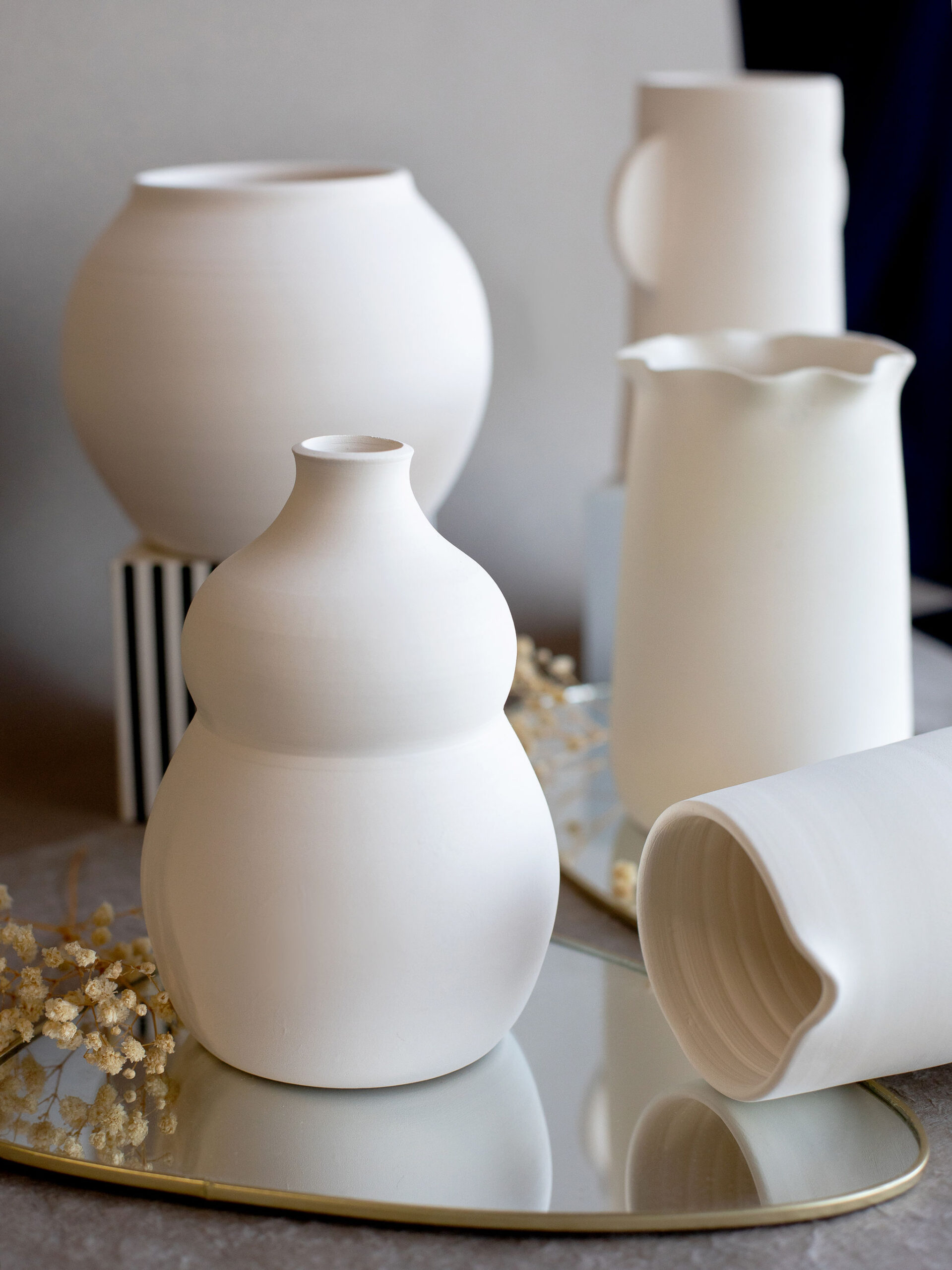 Vases et pots en céramique blanche mate, de formes variées (gourde, sphère, pichet ondulé), sont disposés sur un plateau miroir doré avec des fleurs séchées, sur fond gris.