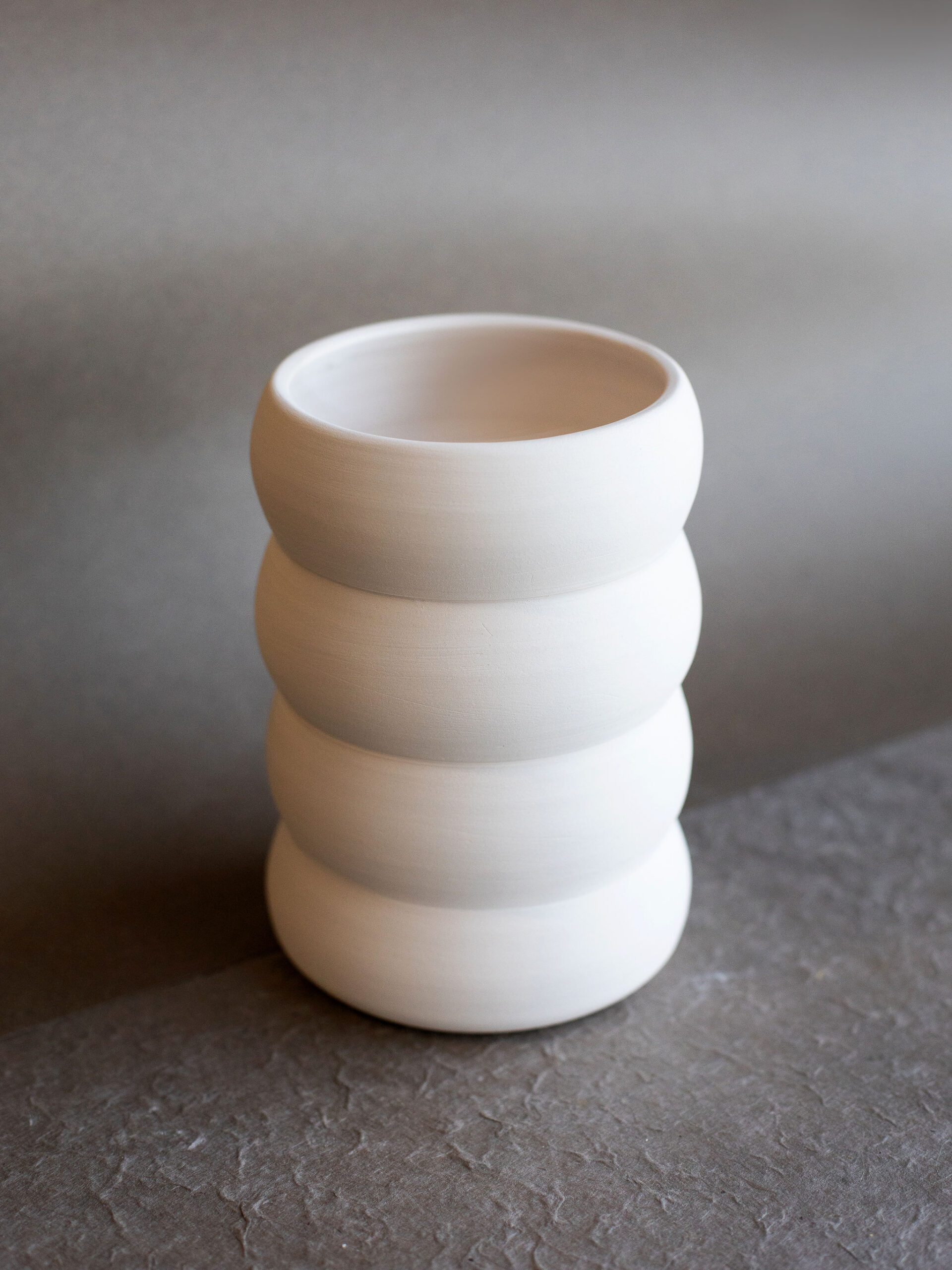 Vase blanc en céramique, à la silhouette empilée de trois anneaux bombés, posé sur une surface texturée gris foncé devant un fond gris clair avec une ombre distincte.