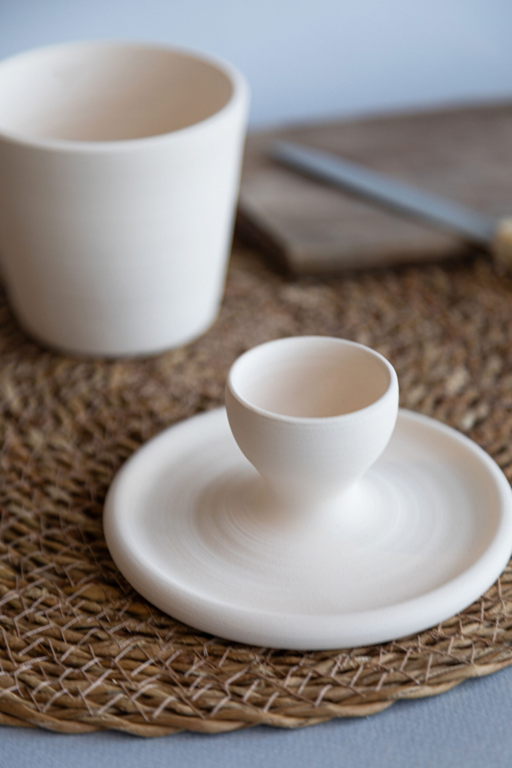 Un coquetier et une assiette en céramique blanche reposent sur un set de table tressé. Un gobelet, une planche à découper et un couteau sont flous à l'arrière-plan.