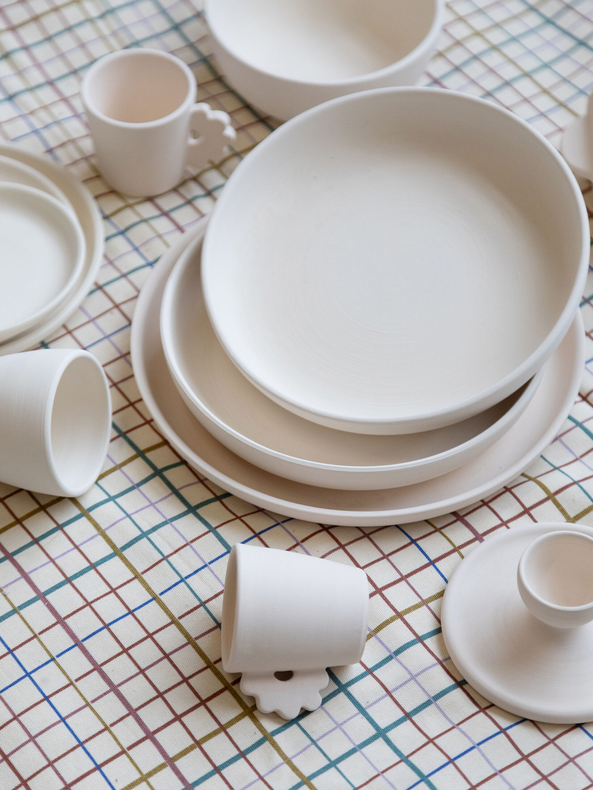 Plusieurs pièces de vaisselle en céramique brute, comprenant des assiettes, bols et tasses, sont éparpillées sur une nappe à carreaux multicolores (verts, marrons, bleus et violets).
