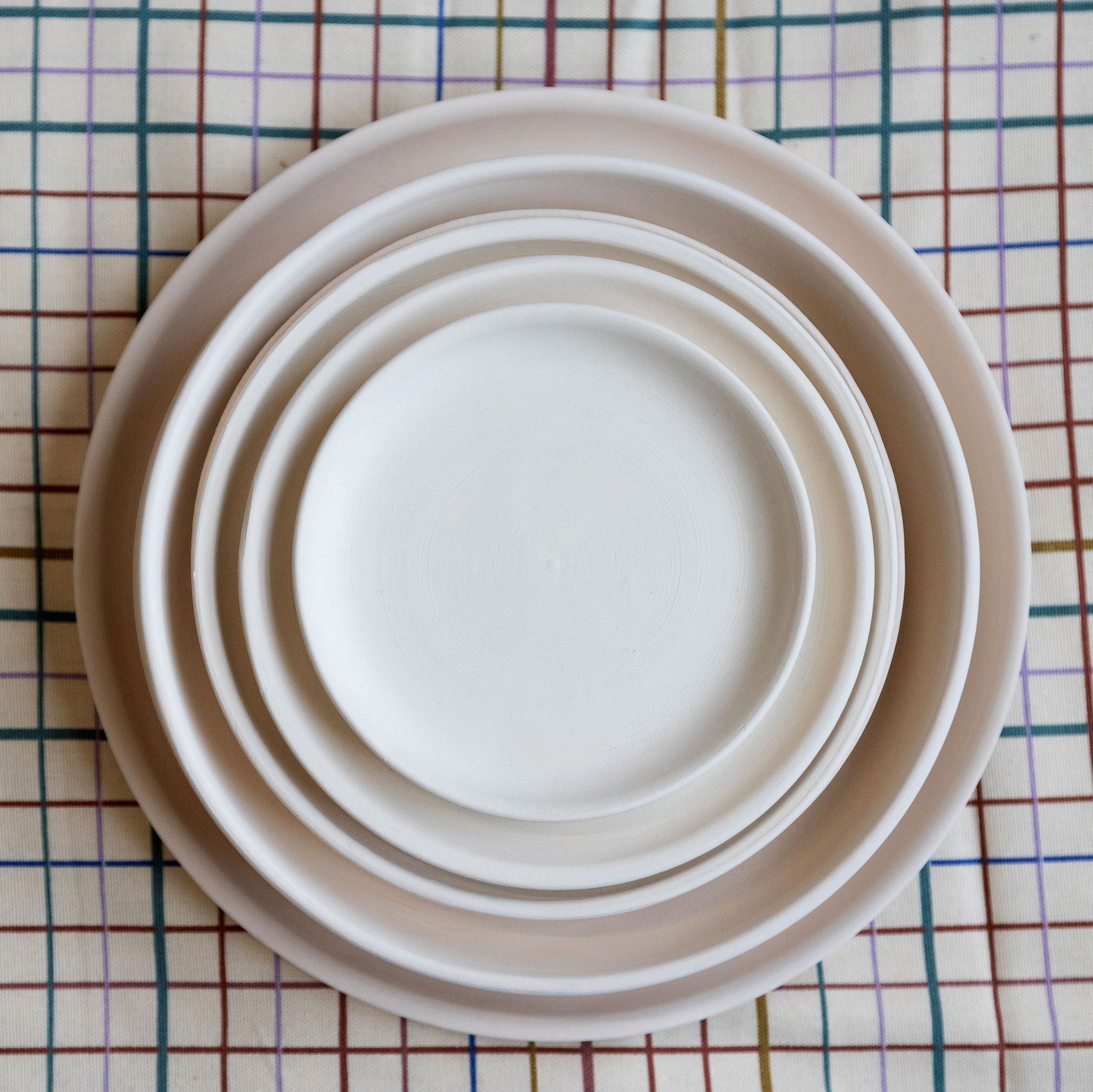 Cinq assiettes rondes blanc cassé sont empilées concentriquement, du plus grand au plus petit, sur une nappe quadrillée aux lignes fines multicolores (marron, bleu, vert, violet).