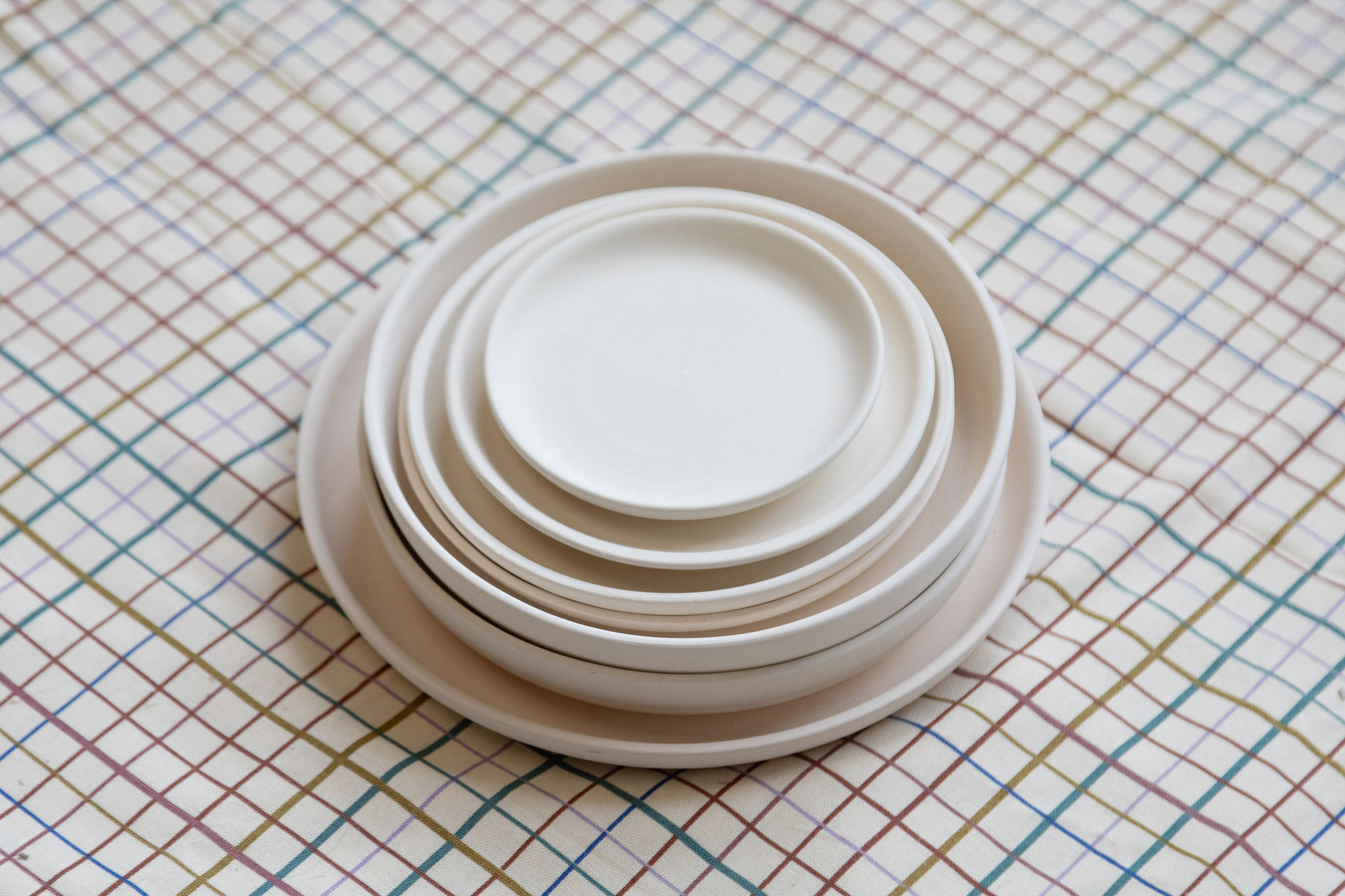 Pile de sept assiettes et bols en céramique blanche, empilés du plus grand au plus petit, posée sur une nappe à carreaux multicolores discrets.