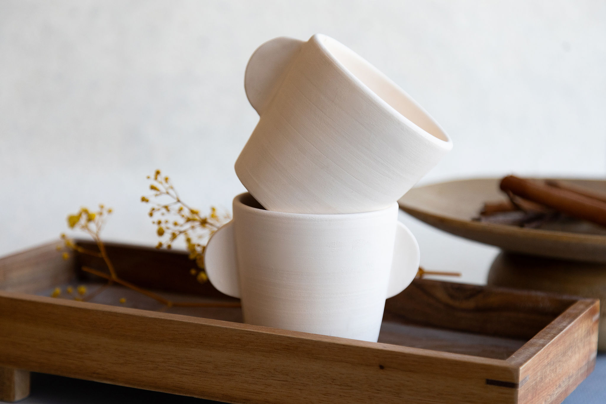 Deux tasses en céramique blanc cassé, à petites anses, sont empilées et inclinées sur un plateau en bois, près de fleurs jaunes séchées et de bâtons de cannelle.