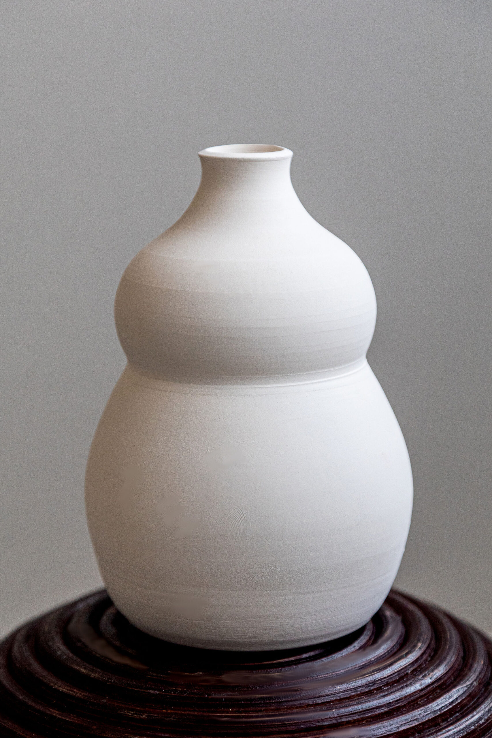 Un vase en céramique blanche mate, de forme gourde, repose sur un socle en bois sculpté foncé. Il se dresse verticalement sur un fond gris clair uniforme.
