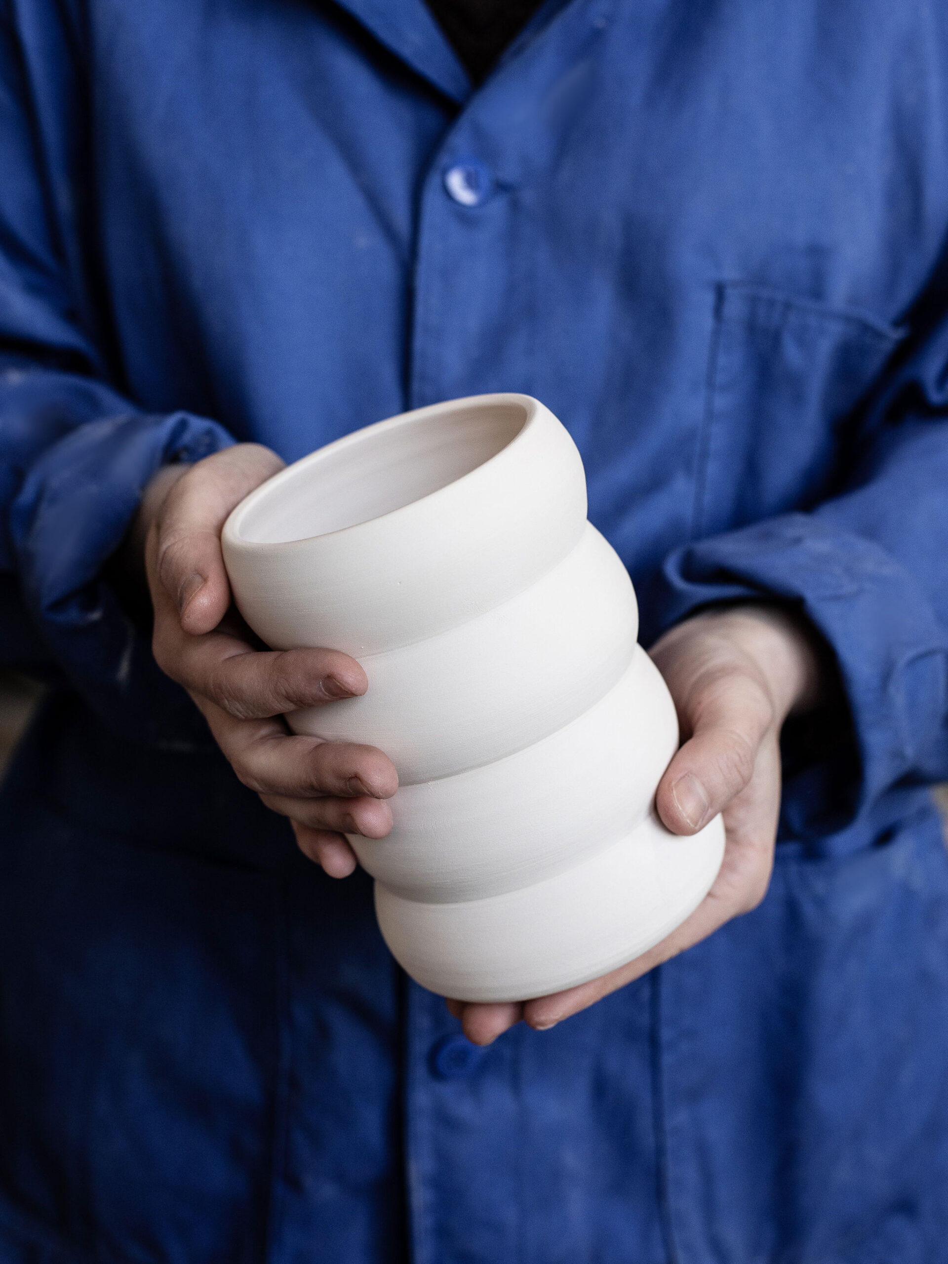 Mains d'une personne tenant fermement un récipient en céramique blanc mat, de forme ondulée avec trois renflements. La personne est vêtue d'une blouse de travail bleue.