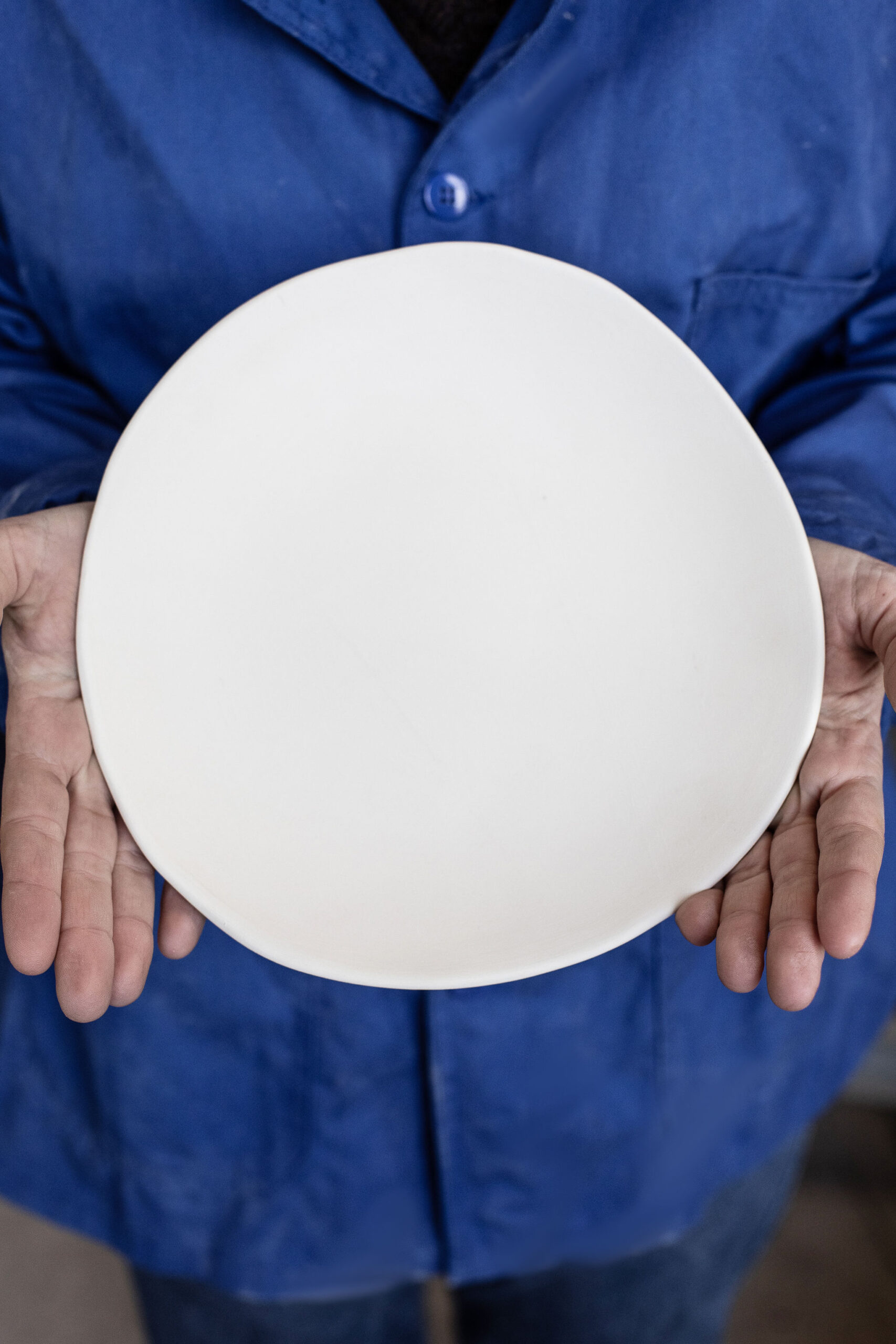 Une personne tient un plat rond et blanc de ses deux mains, devant son corps vêtu d'une blouse de travail bleue.