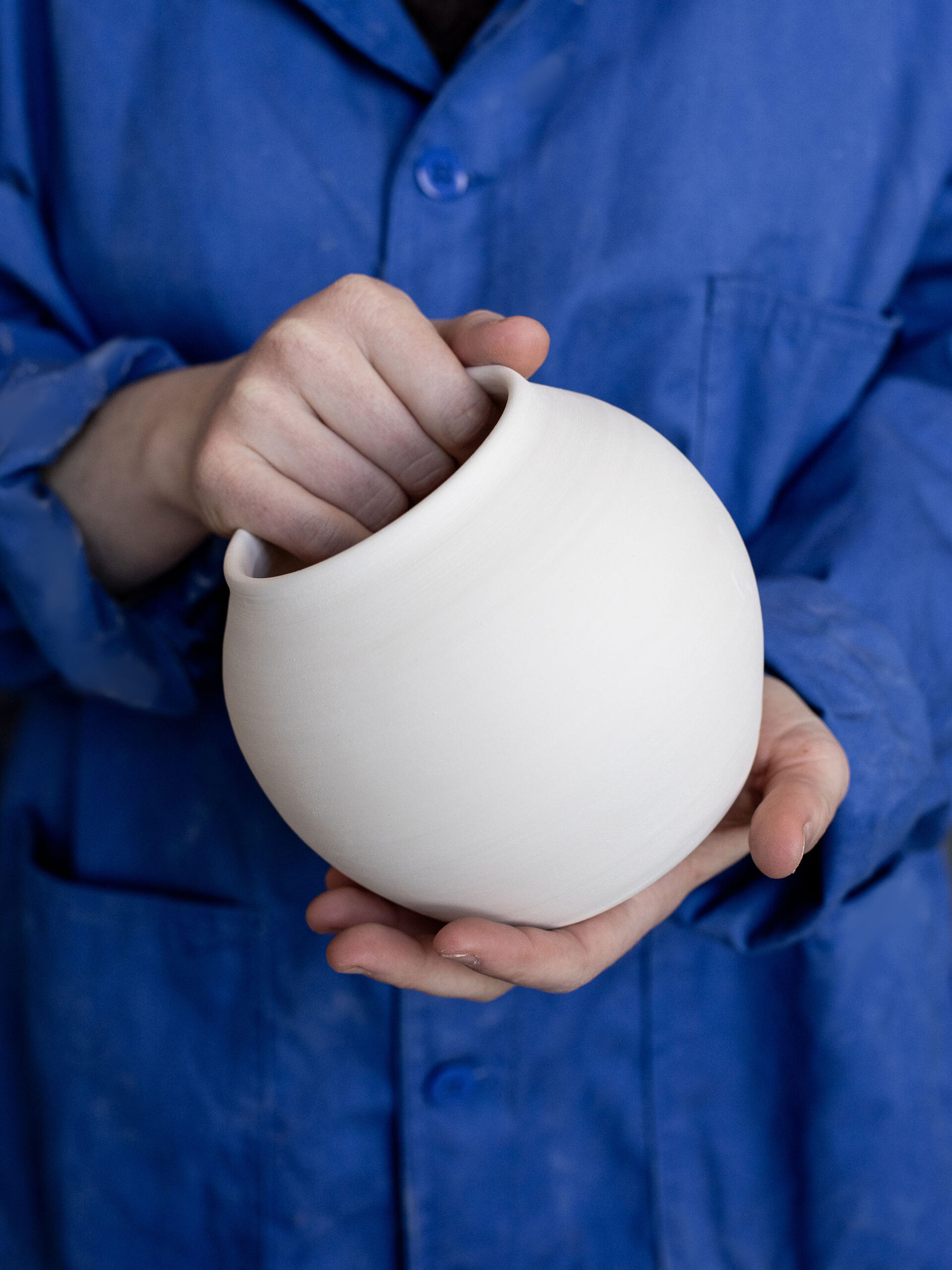 Des mains, vêtues d'une blouse bleue, tiennent une poterie blanche, lisse et sphérique. Une main soutient le bas, l'autre explore son col ouvert.