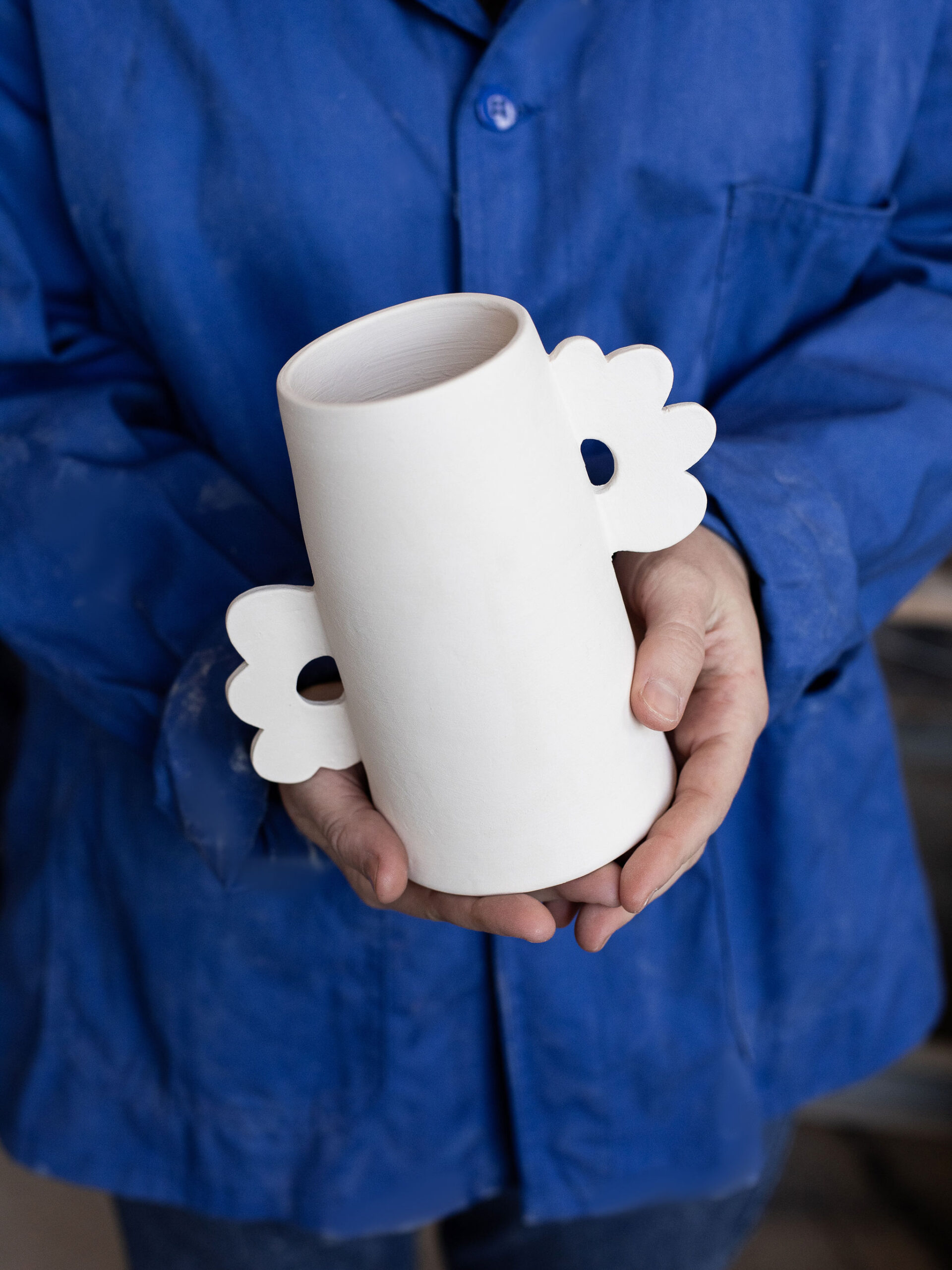 Des mains, vêtues d'une blouse bleue, présentent une tasse cylindrique en céramique blanche. Elle possède deux poignées en forme de fleurs stylisées, une de chaque côté.
