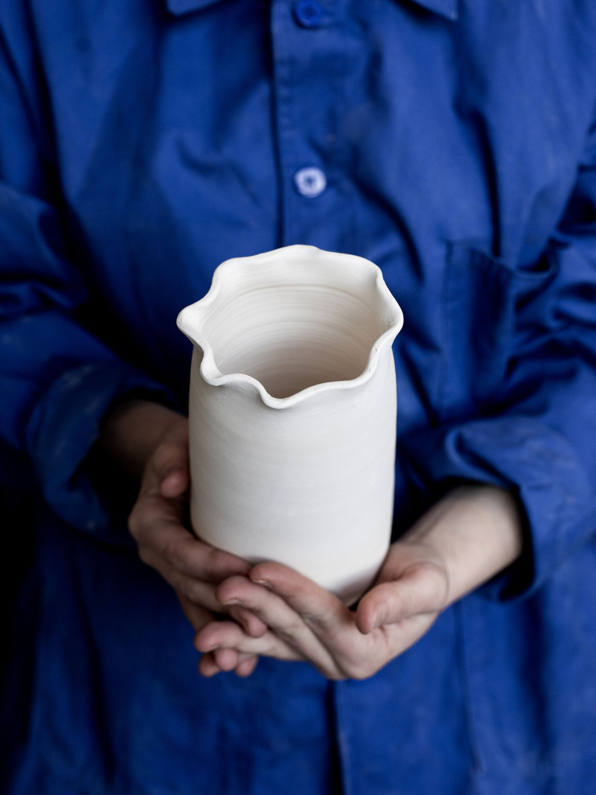 Une personne vêtue d'une veste de travail bleue tient à deux mains un pot en céramique blanc brut à l'ouverture ondulée.