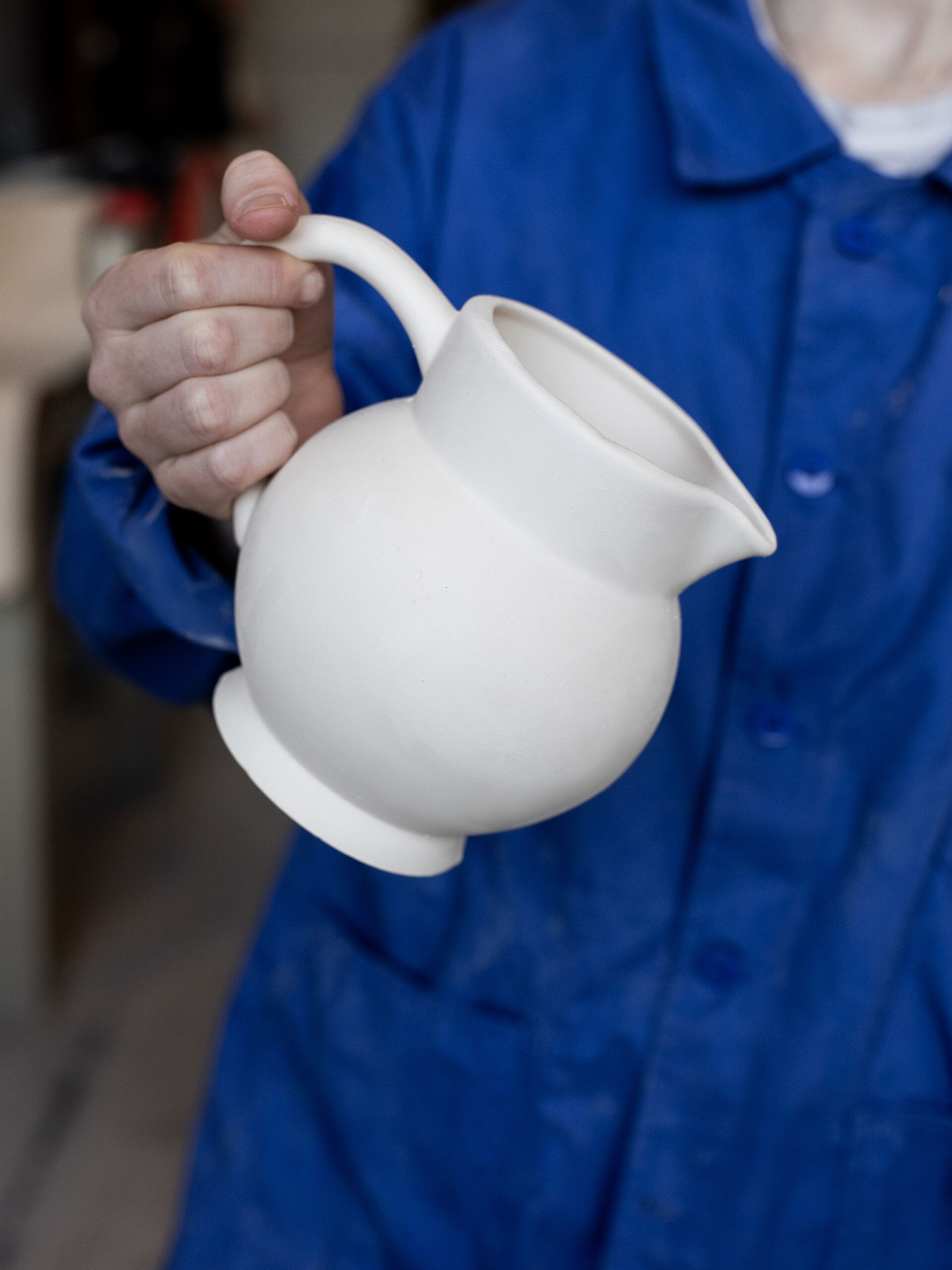 Une main tient une cruche en céramique blanche mate, à col évasé et bec verseur. La personne porte une veste de travail bleue dans un atelier.