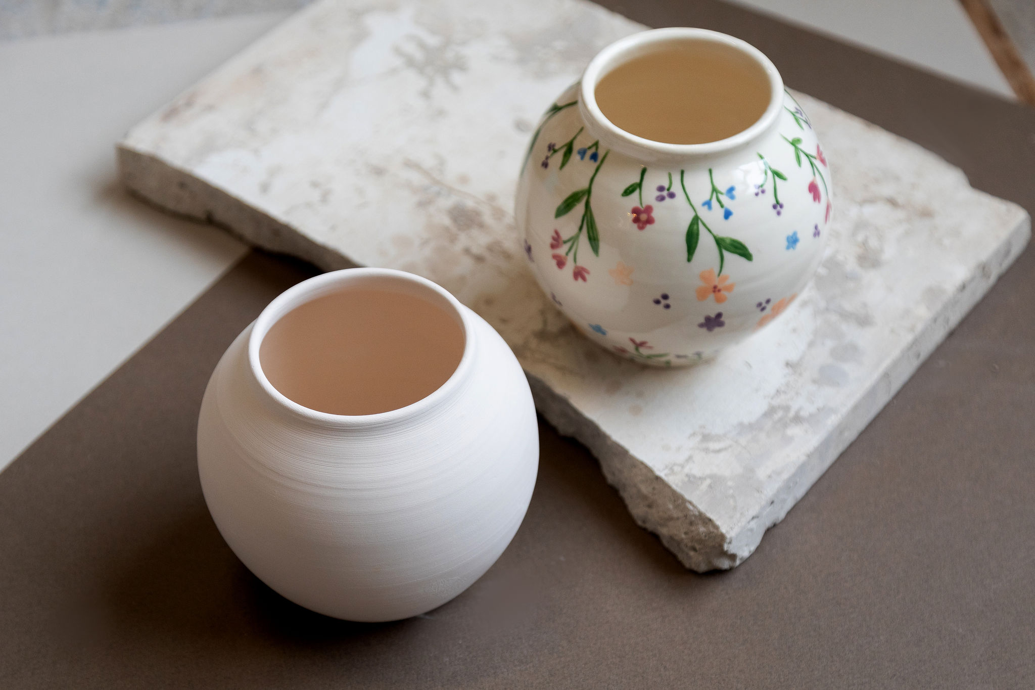 Deux petits pots ronds en céramique, l'un blanc uni, l'autre à motifs floraux colorés, sont posés côte à côte sur une surface claire.
