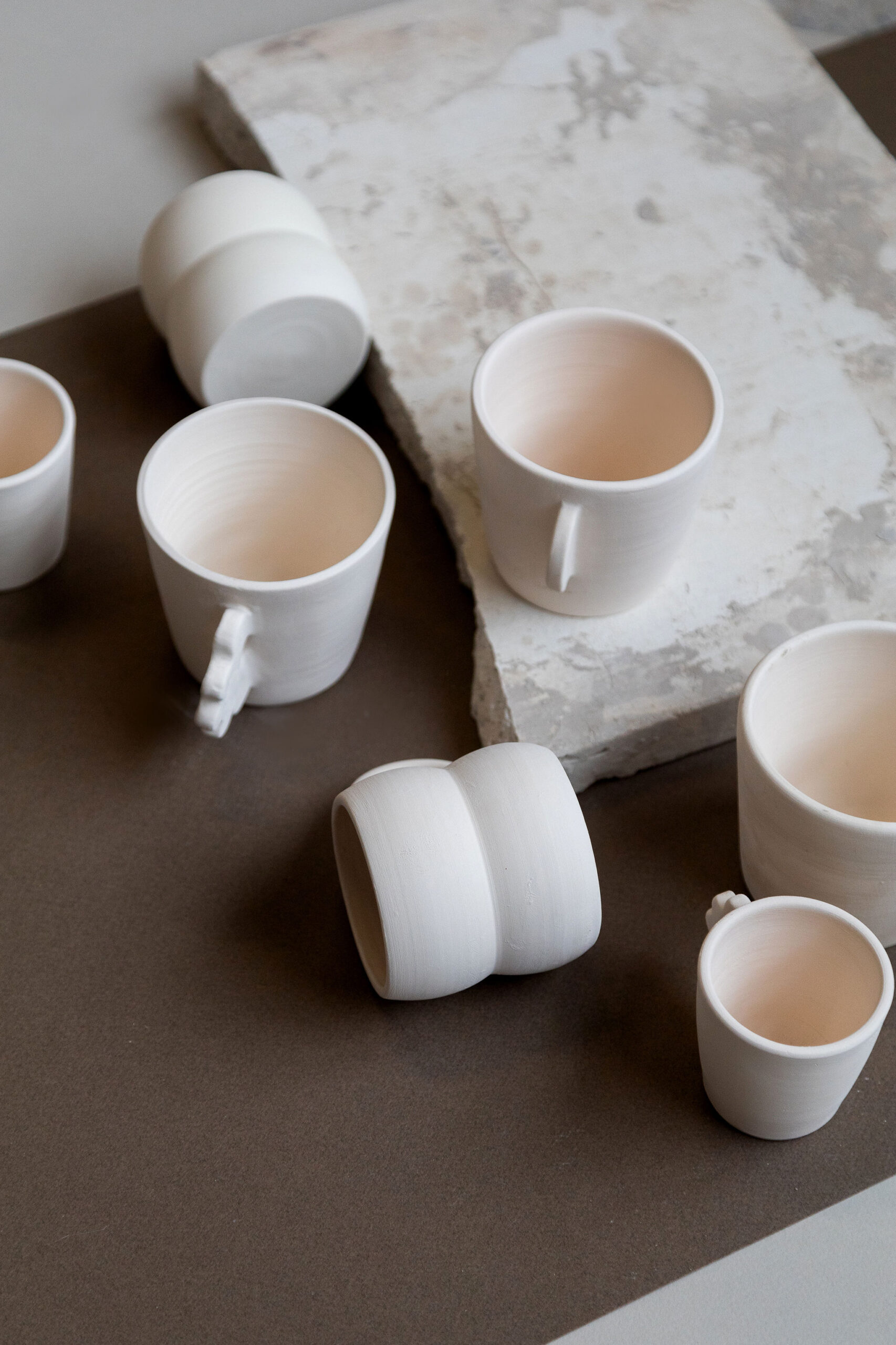 Des tasses et verres en céramique blanche mate, certains avec anse, sont disposés de manière informelle sur une surface bicolore marron et beige clair, avec des blocs de pierre.