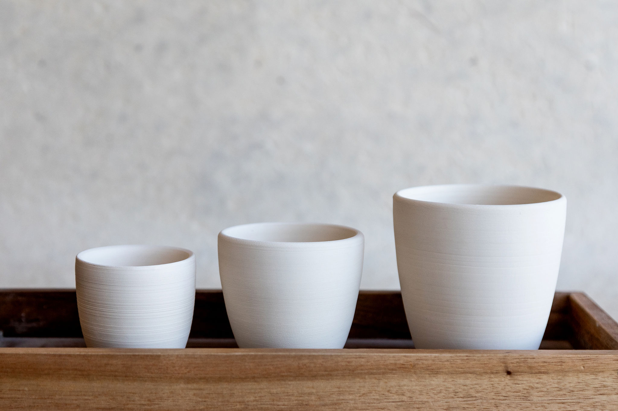 Trois tasses en céramique blanche sont alignées par taille décroissante sur une étagère en bois. Elles se détachent sur un fond clair et flou.