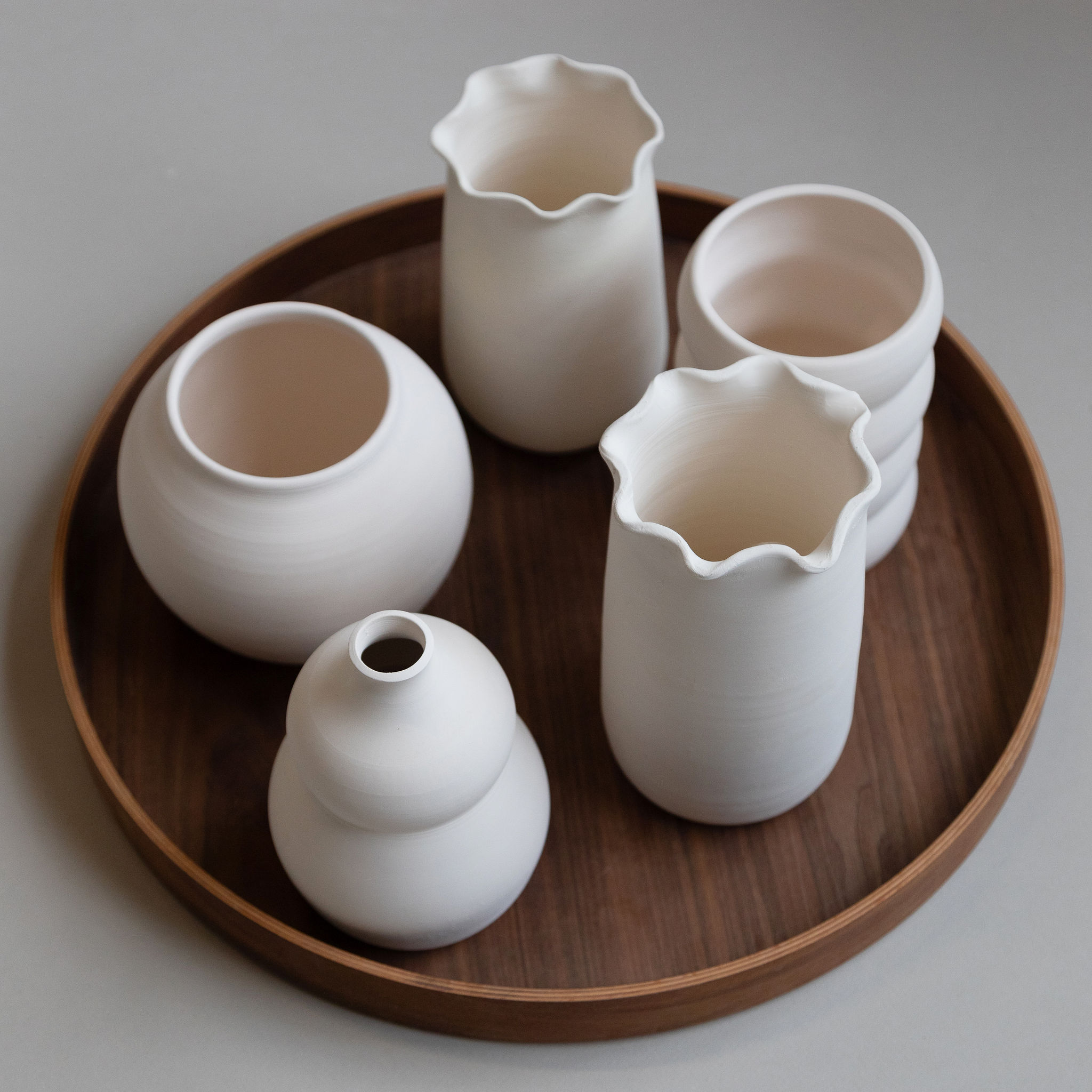 Cinq vases et pots en céramique blancs mats, aux formes variées et bouches ondulées ou lisses, sont disposés sur un plateau rond en bois foncé, posé sur une surface grise.