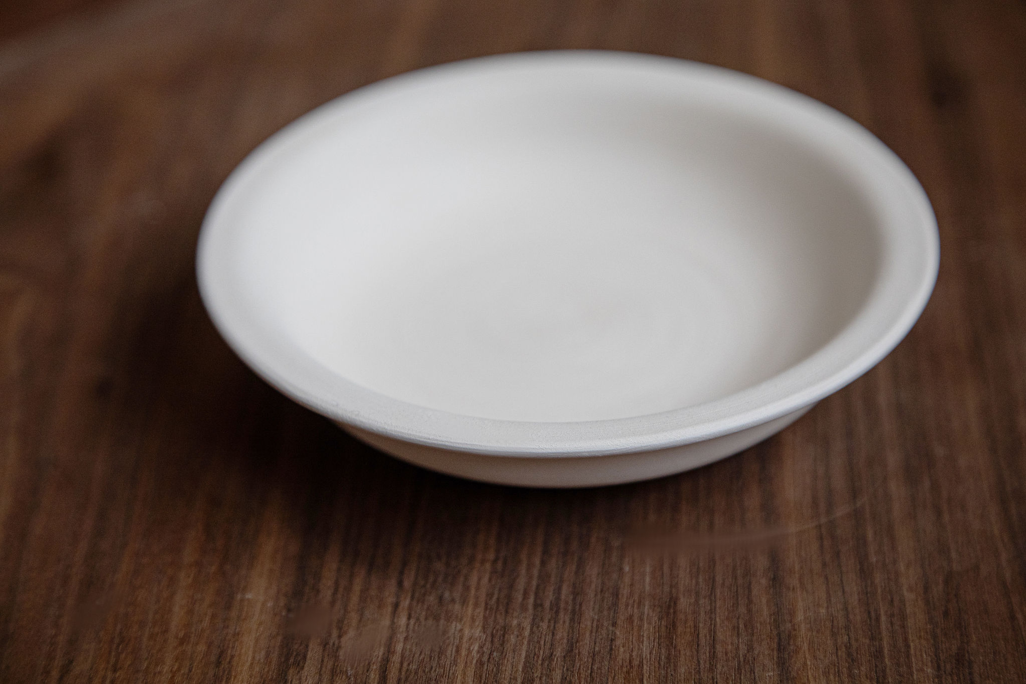 Une petite assiette ovale en céramique blanche, vide et aux bords évasés, repose légèrement inclinée sur une surface en bois brun foncé avec un grain visible.