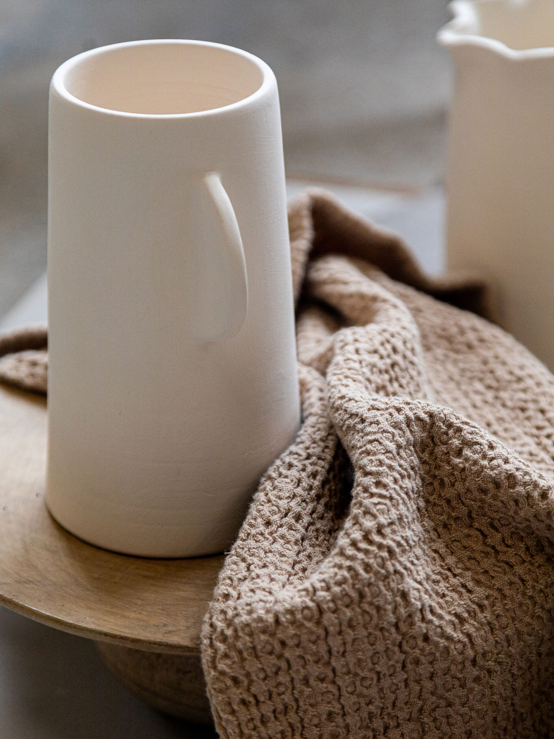 Tasse haute en céramique beige clair, légèrement texturée, repose sur une surface en bois clair. Un tissu froissé, d'une teinte brun-beige, est posé à ses côtés.