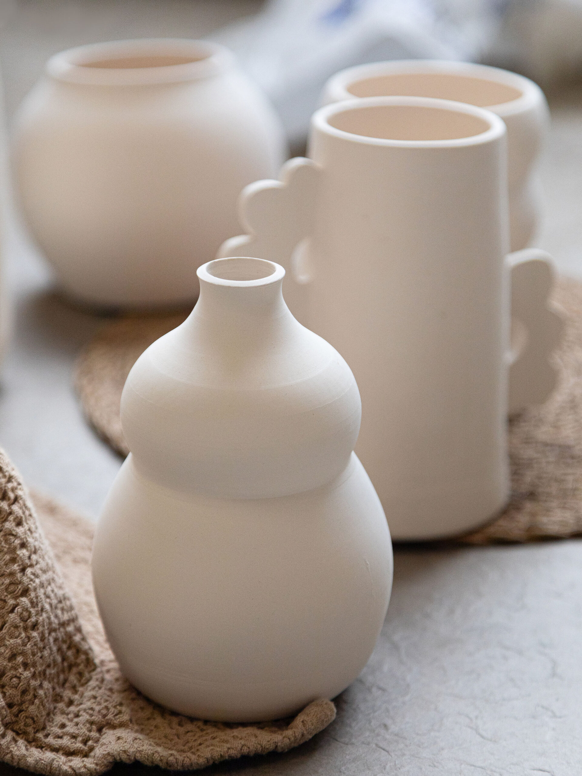 Diverses pièces de poterie blanche mate, notamment un vase en forme de gourde et des tasses, sont disposées sur une table claire avec des napperons texturés, prêtes à être décorées.