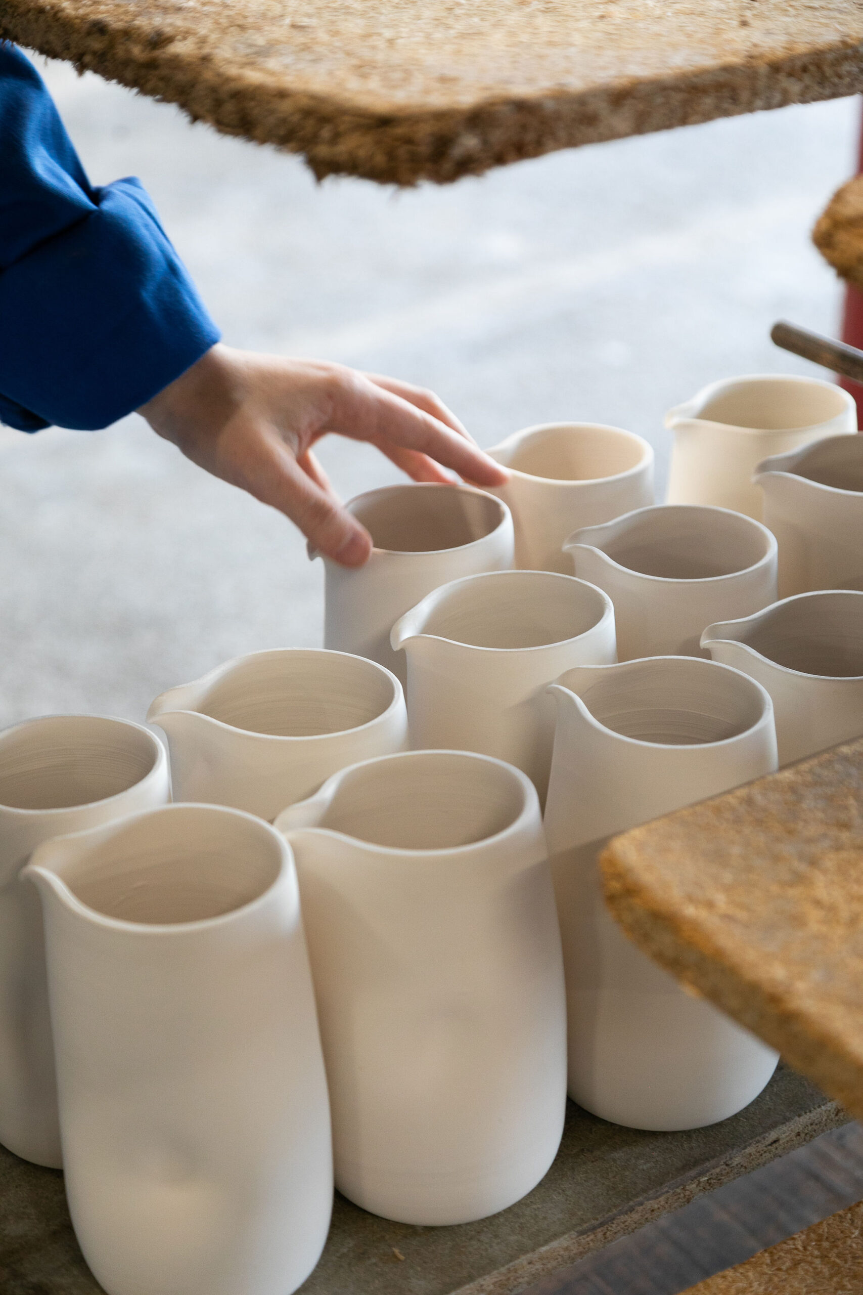 Une main touche une petite carafe en céramique blanche, parmi plusieurs récipients identiques alignés sur une surface grise, sous une étagère en bois.