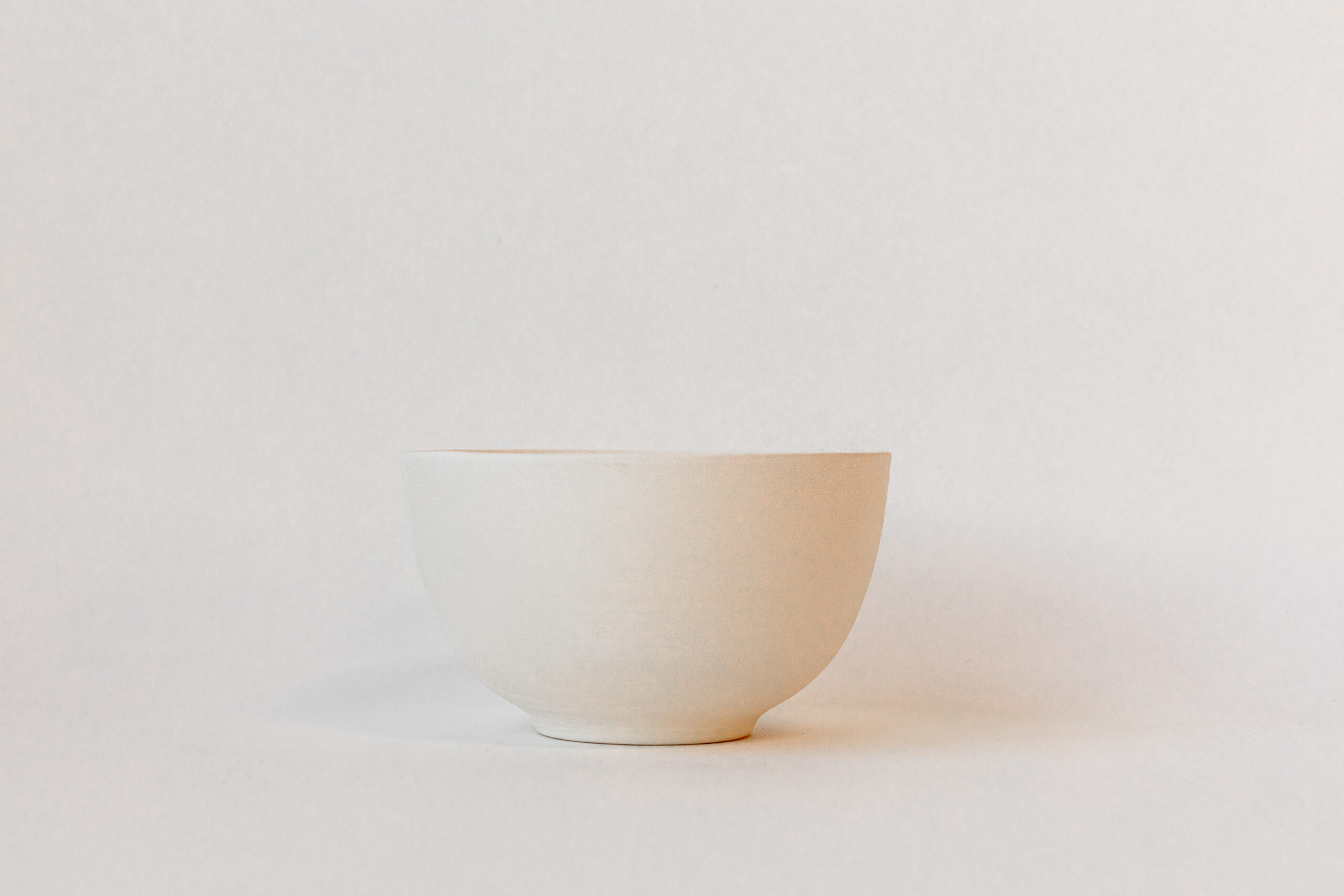 Un bol épuré en céramique, de couleur blanc cassé et vide, est posé sur une surface claire devant un fond uni et lumineux.