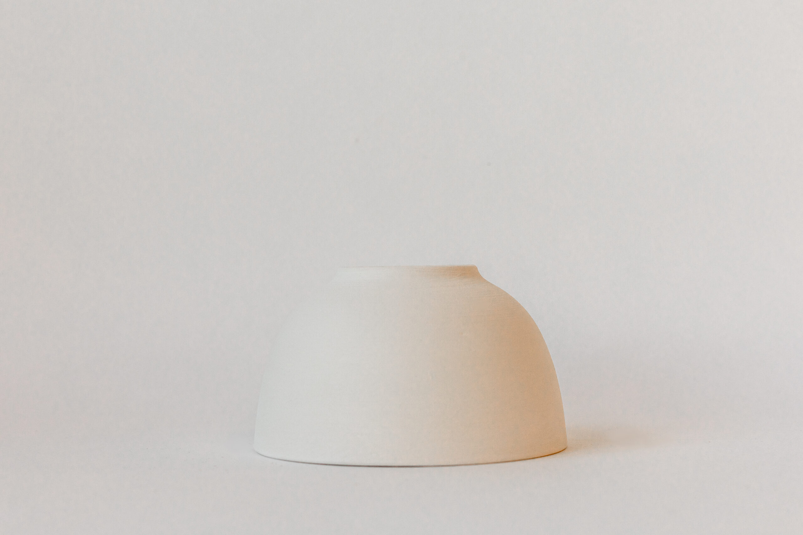 Un petit bol en céramique mate, blanc cassé, de forme arrondie, est posé sur une surface claire. Le fond est uni et minimaliste.