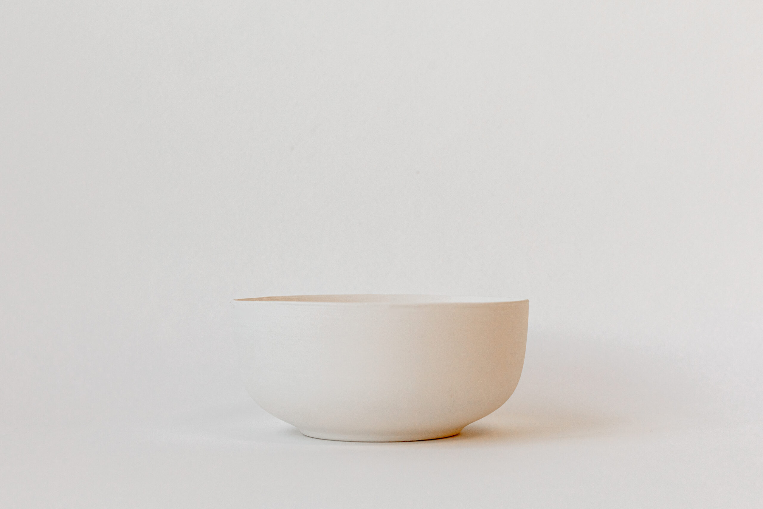 Un bol simple, de couleur blanc cassé, est posé au centre d'une surface plane devant un fond uni de la même teinte claire.
