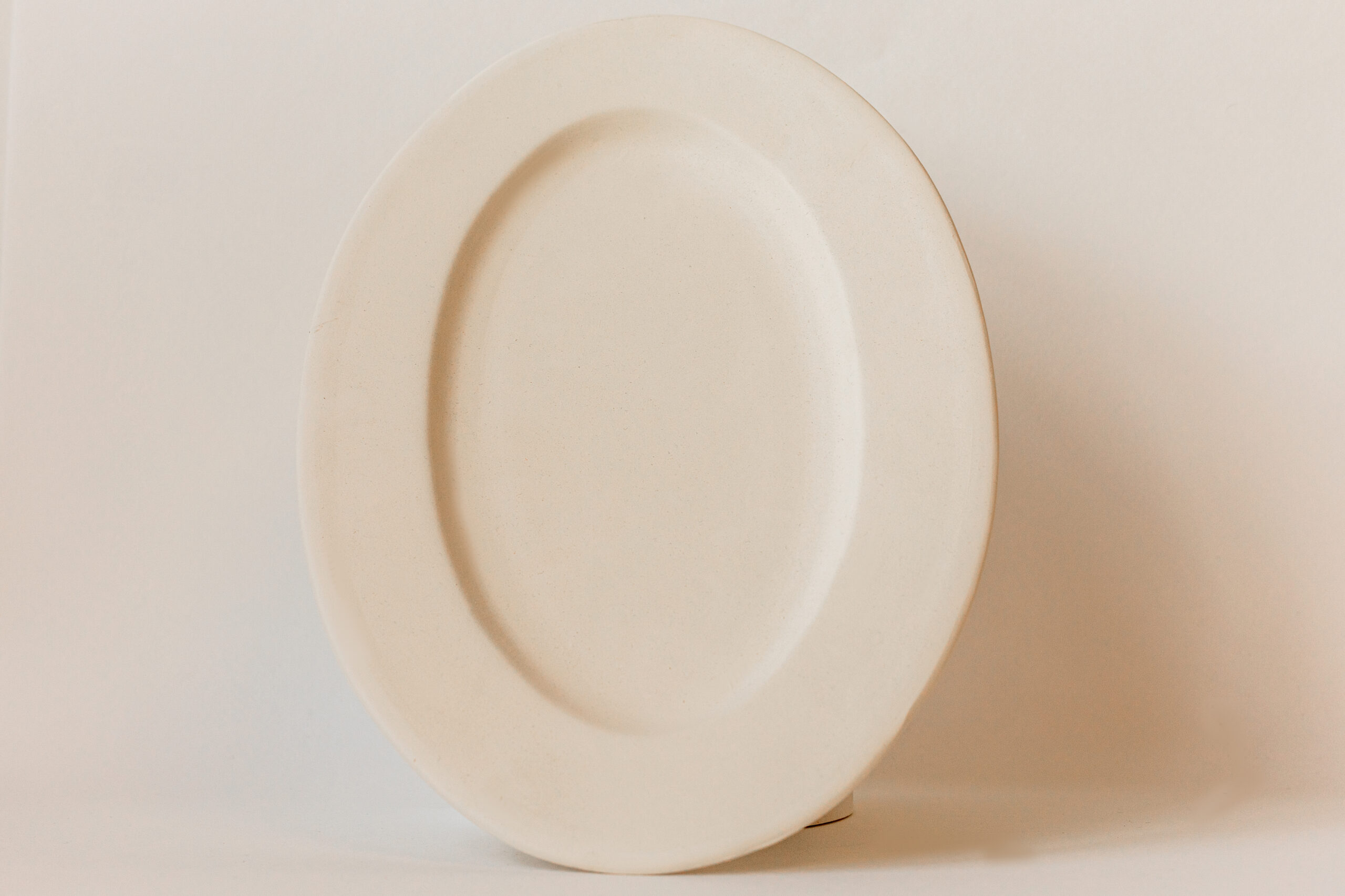 Une assiette en céramique, de couleur crème et vide, se dresse légèrement inclinée sur un fond clair et uni, présentant un léger rebord.