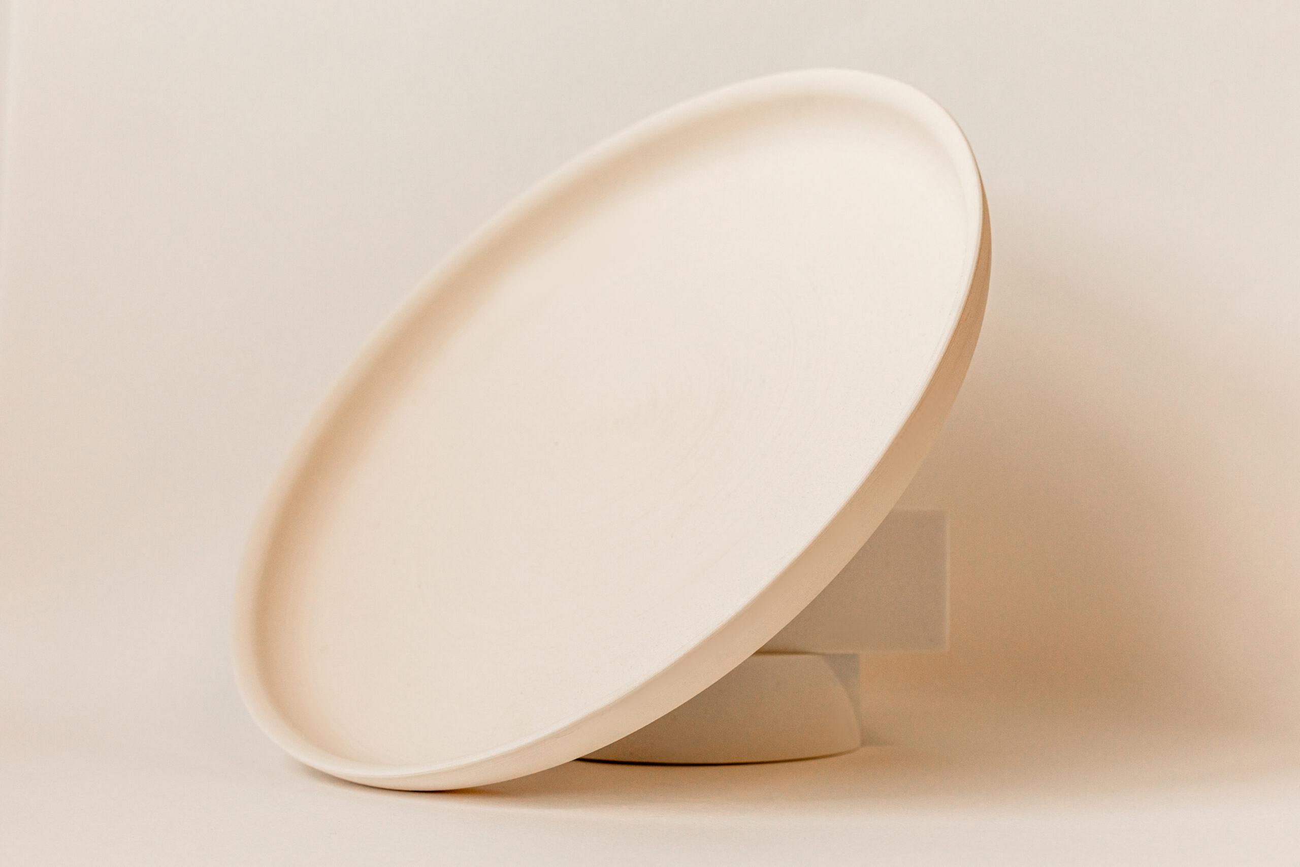Une assiette ovale en céramique de couleur crème, légèrement inclinée et posée verticalement, sur un fond uni et lumineux. Style minimaliste et épuré.
