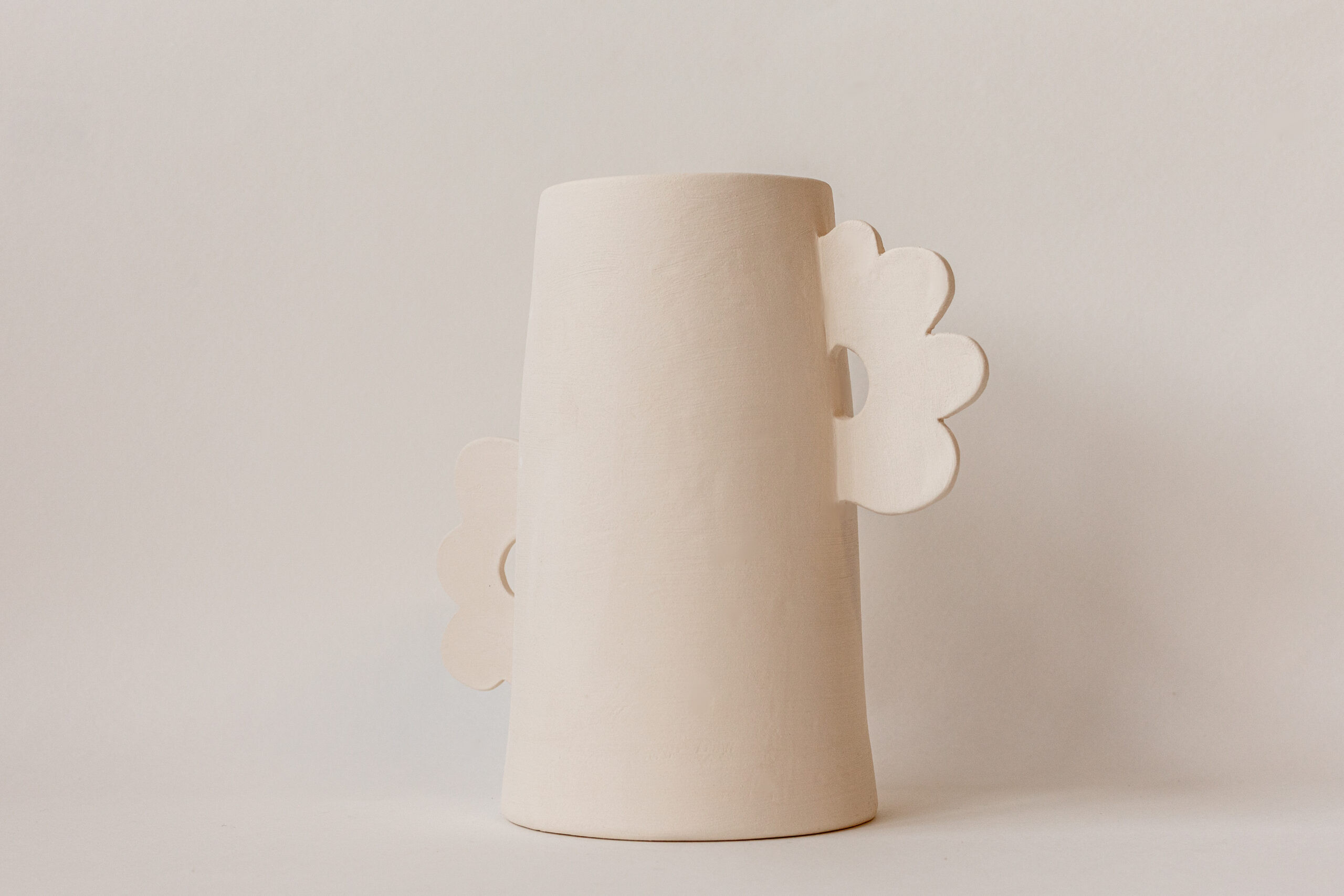Vase cylindrique conique beige clair avec une anse ondulée et un élément décoratif floral similaire sur le bord opposé, posé sur un fond uni et lumineux.