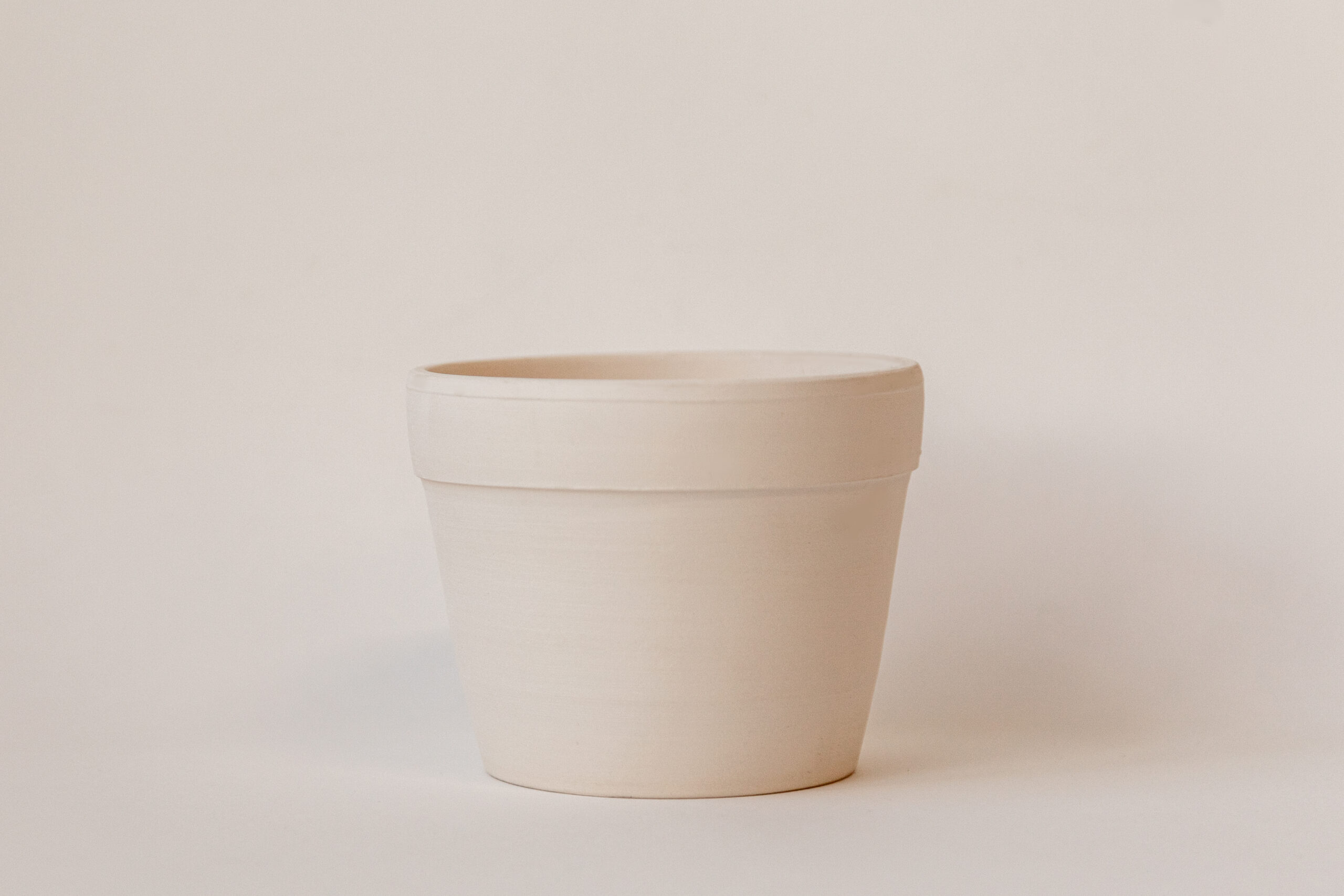 Un petit pot de fleurs cylindrique, vide et de couleur beige clair, est posé frontalement sur un fond uni et immaculé. Son aspect est simple et fonctionnel.