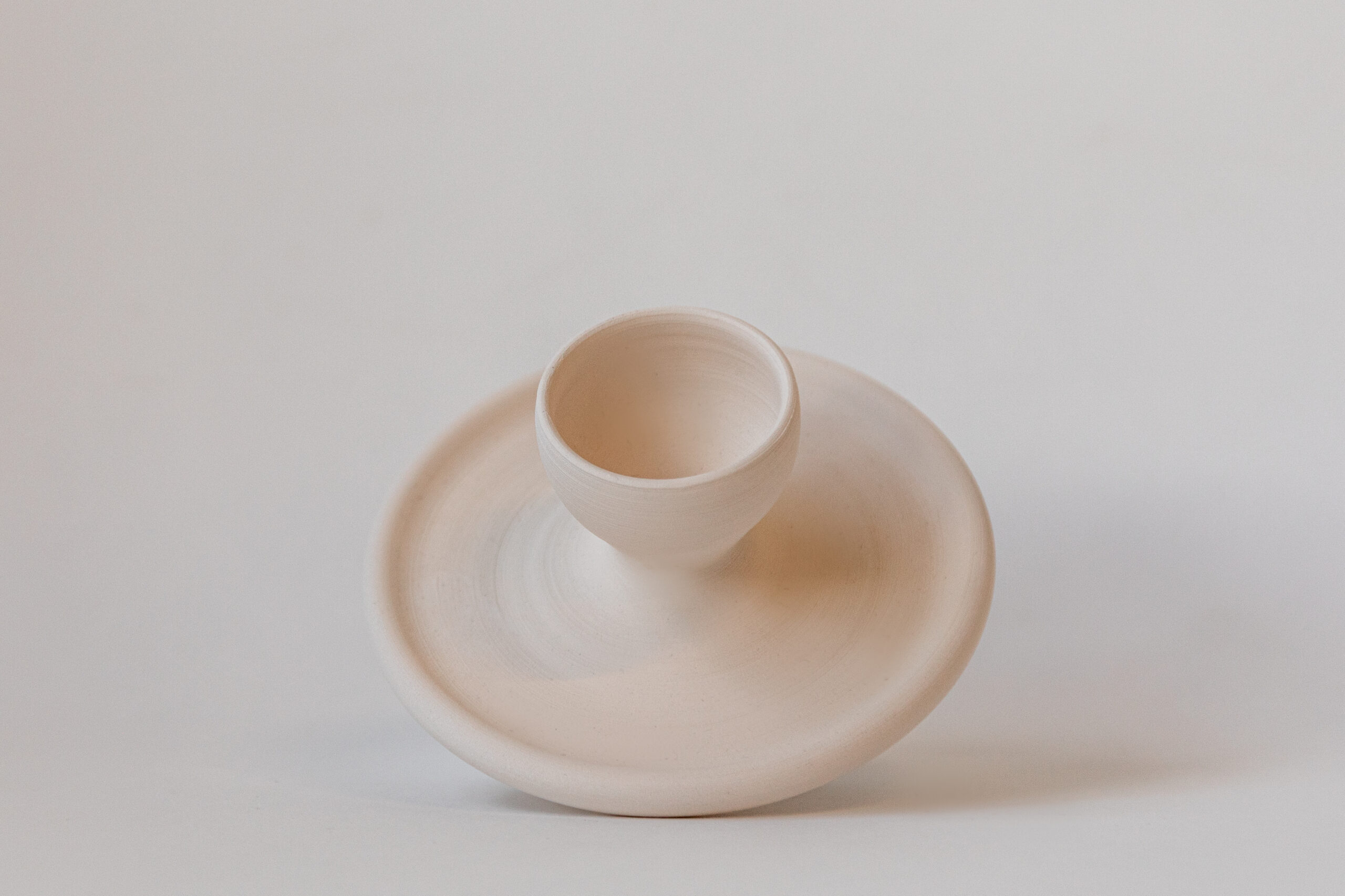 Une sculpture organique en céramique claire présente une base plate d'où s'élève un col tubulaire asymétrique. L'objet repose sur un fond blanc uni.