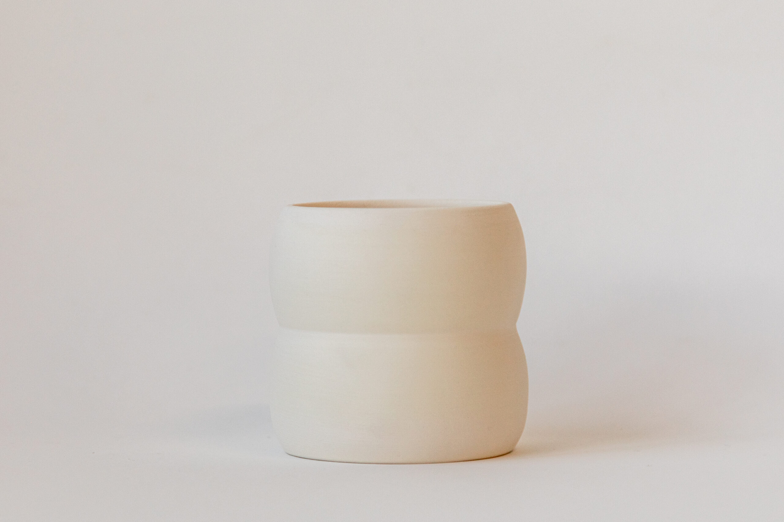 Un gobelet ou petit vase en céramique crème, de forme cylindrique avec une légère courbure au centre, est posé sur un fond uni et clair.