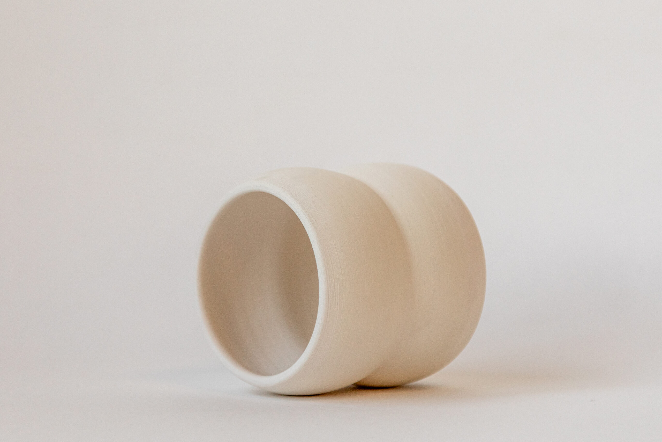 Une tasse cylindrique en céramique claire repose inclinée sur le côté, ouverture vers la gauche, sur un fond blanc uni et minimaliste.