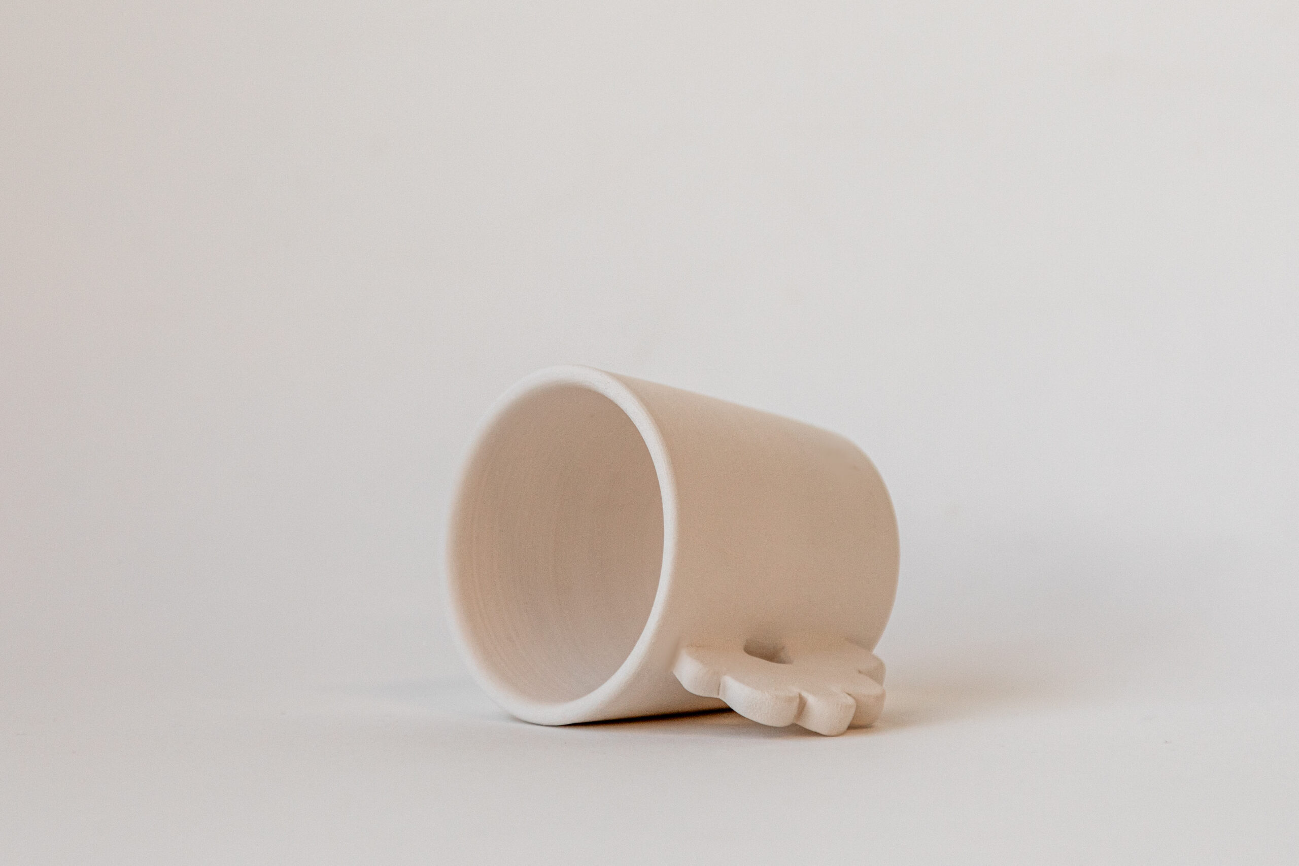 Une tasse beige pâle est couchée sur le côté. À côté, un petit objet en forme de fleur de la même couleur repose sur un fond clair uni.