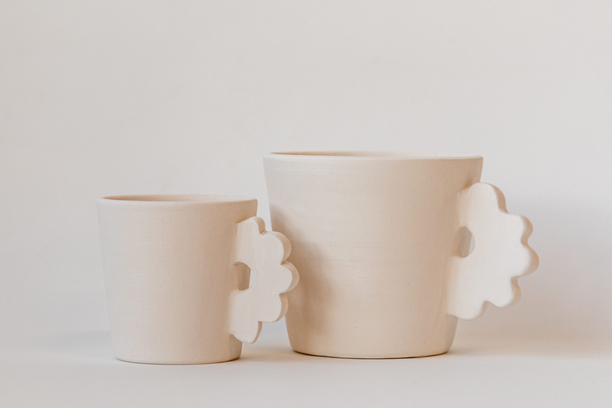 Deux tasses en céramique beige mat, l'une plus grande que l'autre, avec des poignées découpées, reposent sur un fond clair uni.