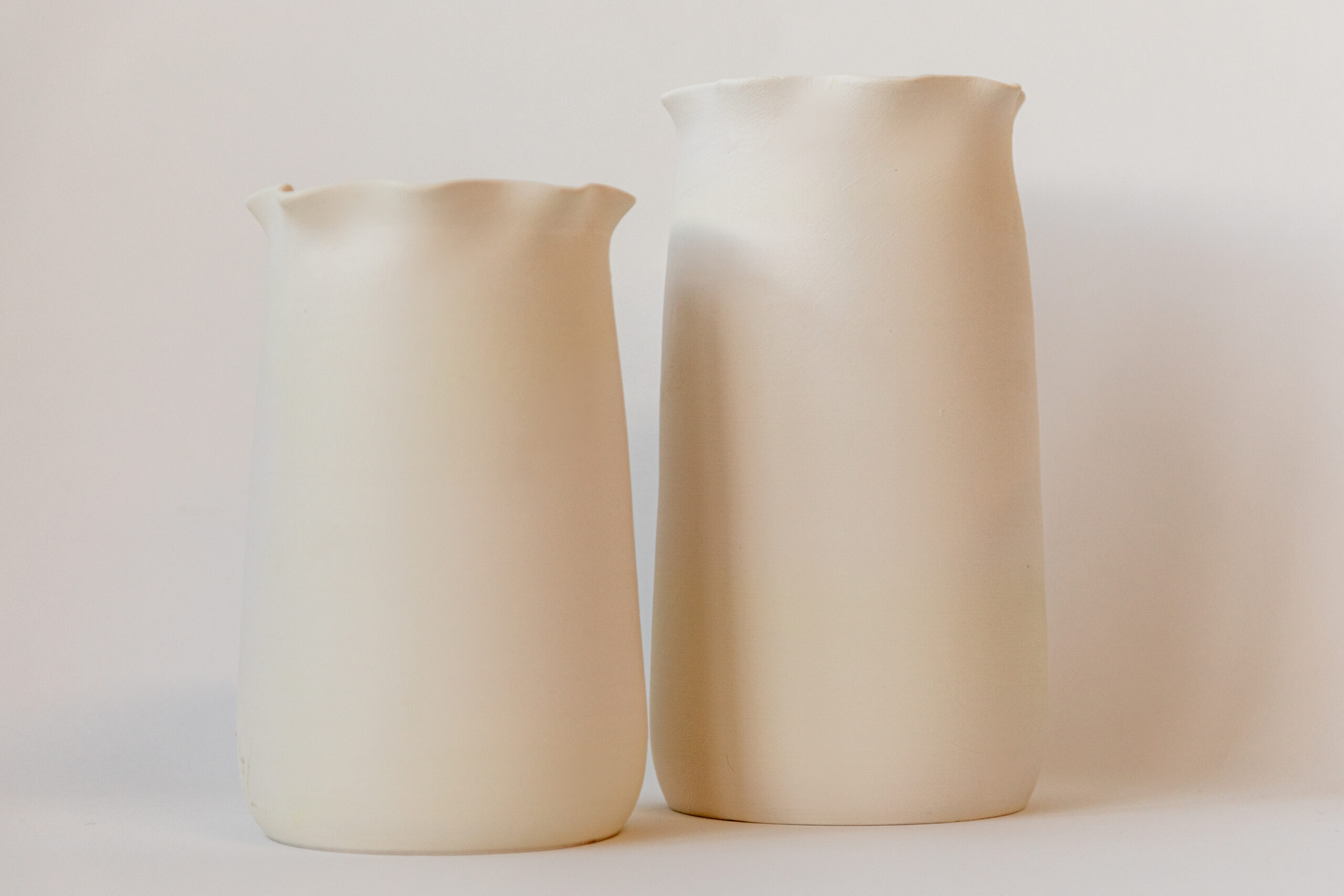 Deux pots cylindriques blancs, l'un plus court, se dressent sur un fond uni clair. Leurs rebords supérieurs sont ondulés, apportant une touche décorative.