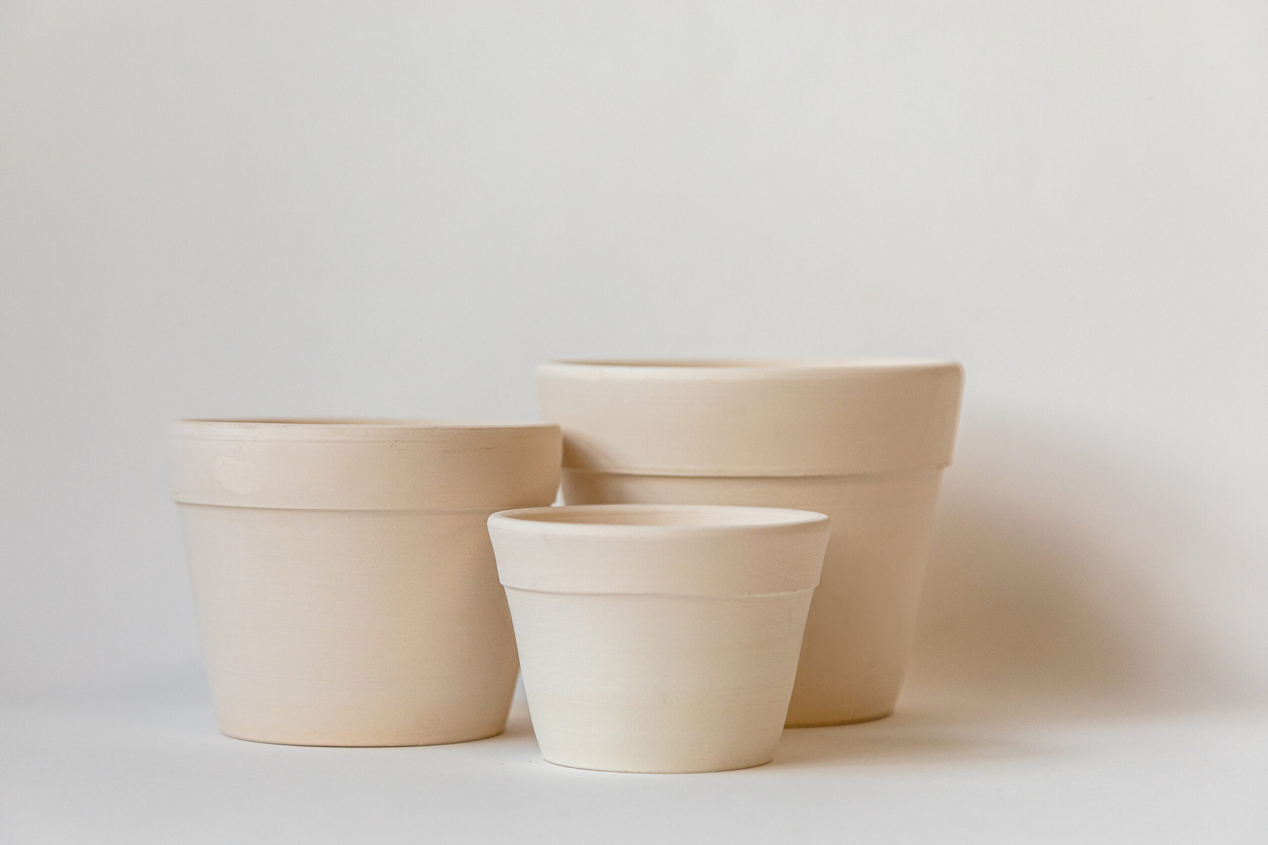 Trois pots en céramique clairs, vides et de tailles variées, se dressent côte à côte sur un fond uni et lumineux.